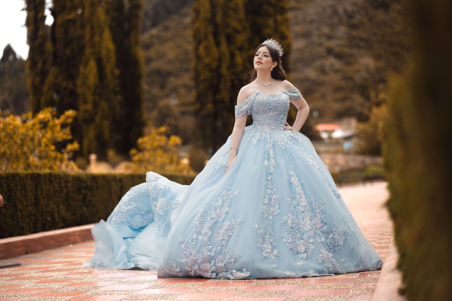 Quinceañera con vestido azul princesa bordado y tiara posa elegante en sendero jardín; fondo árboles altos.