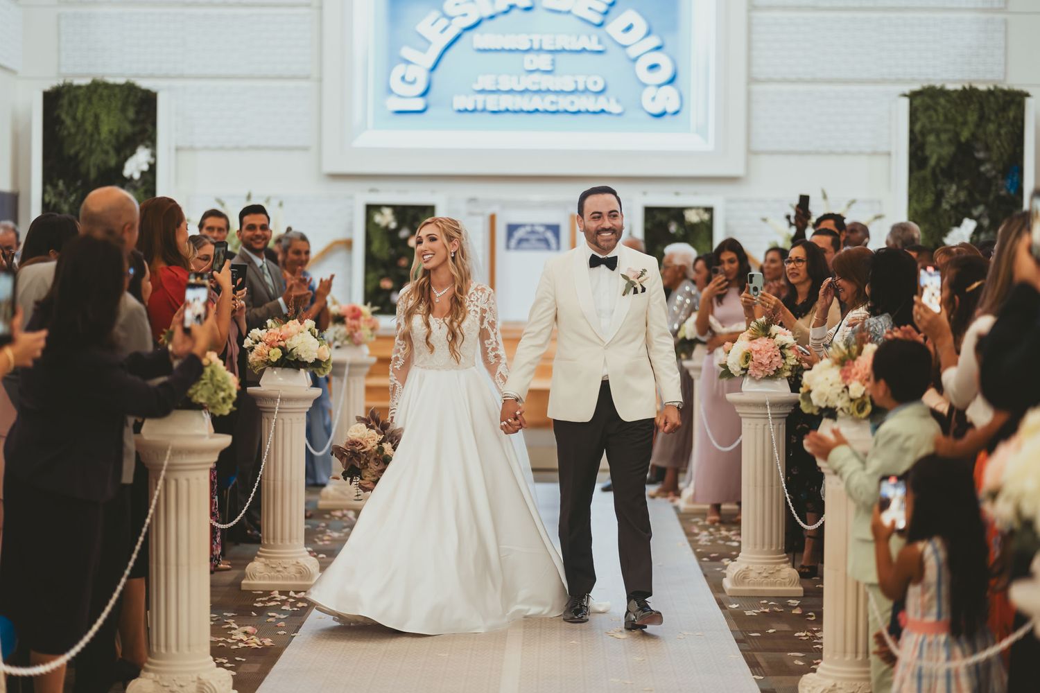 Novios recién casados caminando sonrientes por pasillo de iglesia, invitados aplauden y celebran su boda. Boda IDMJI