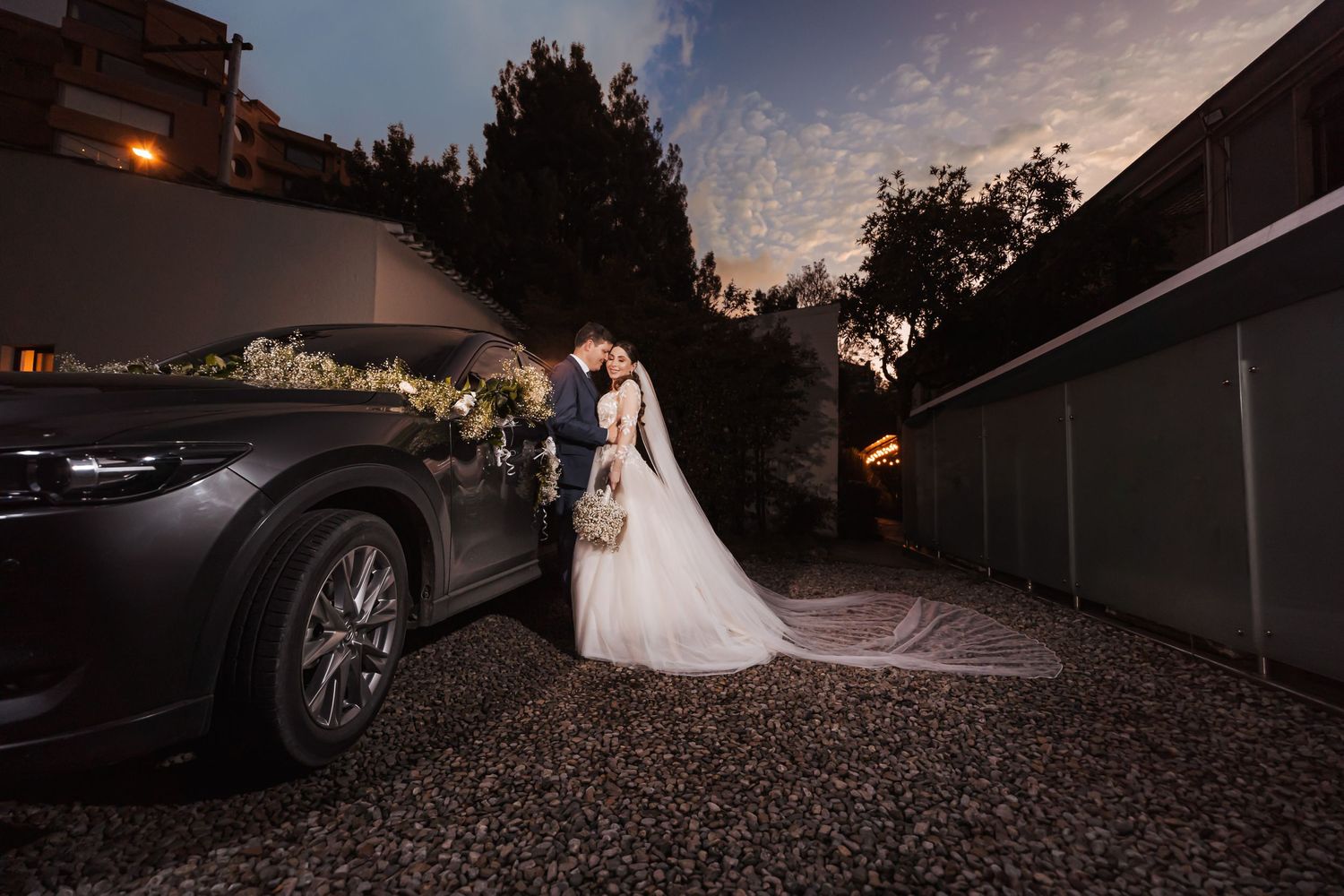 Novios se abrazan junto a coche de boda decorado con flores, al atardecer. Foto romántica de su gran día y velo de la novia
