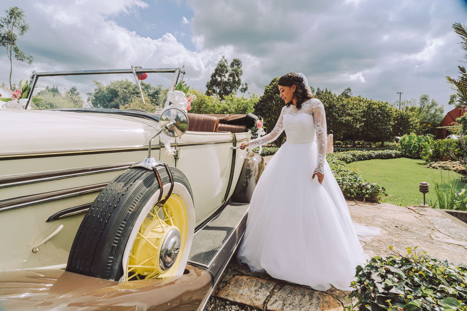 Novia posando junto a un coche antiguo en el jardín de su boda.