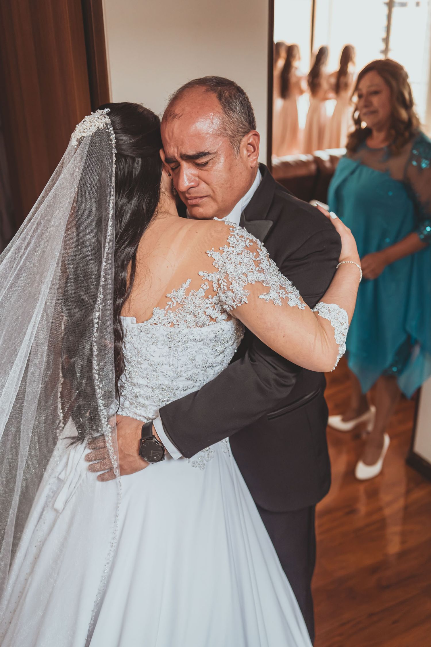 Novia (vestido encaje) abraza con emoción a su padre (esmoquin), quien llora; momento tierno boda.
