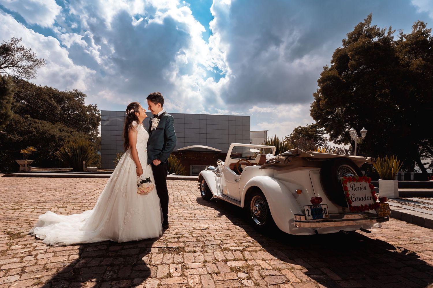 Novios se miran y sonríen emotivamente junto a coche descapotable blanco vintage, bajo cielo nublado. Boda romántica.
