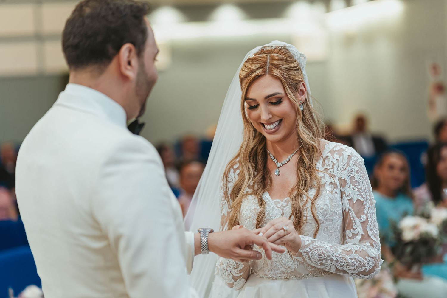 Novio (traje blanco) coloca anillo en dedo de novia sonriente (vestido encaje, velo) durante ceremonia boda.