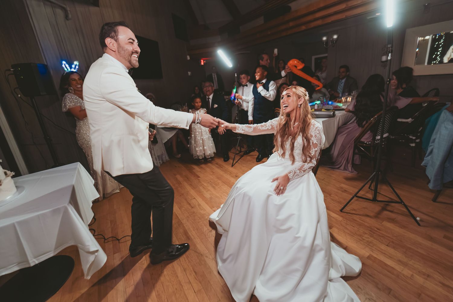 Novia y novio (chaqueta blanca) bailan sonrientes en recepción de boda; invitados al fondo.