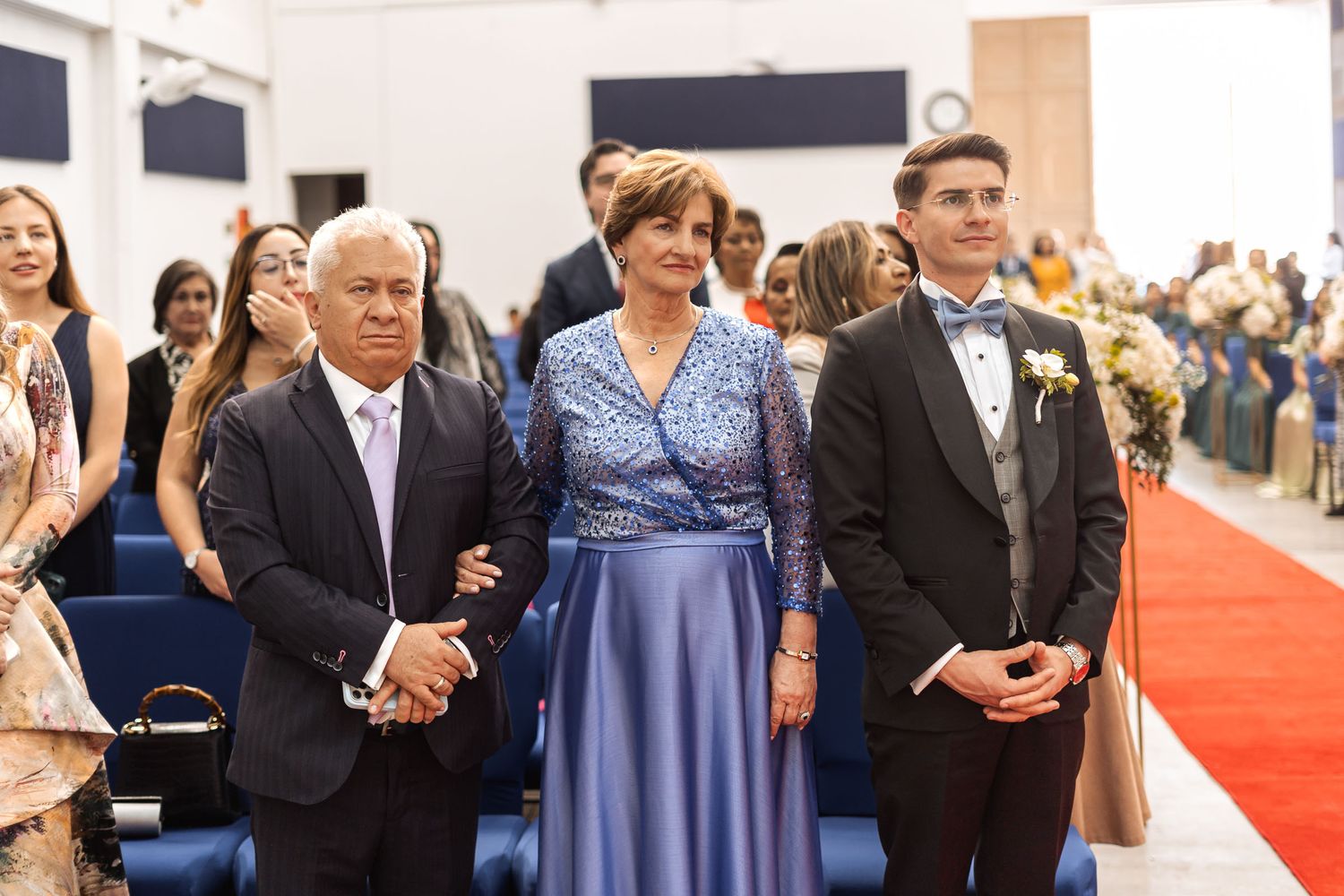 Novio con esmoquin junto a sus padres (o familiares cercanos) durante ceremonia de boda en iglesia; madre viste de azul.