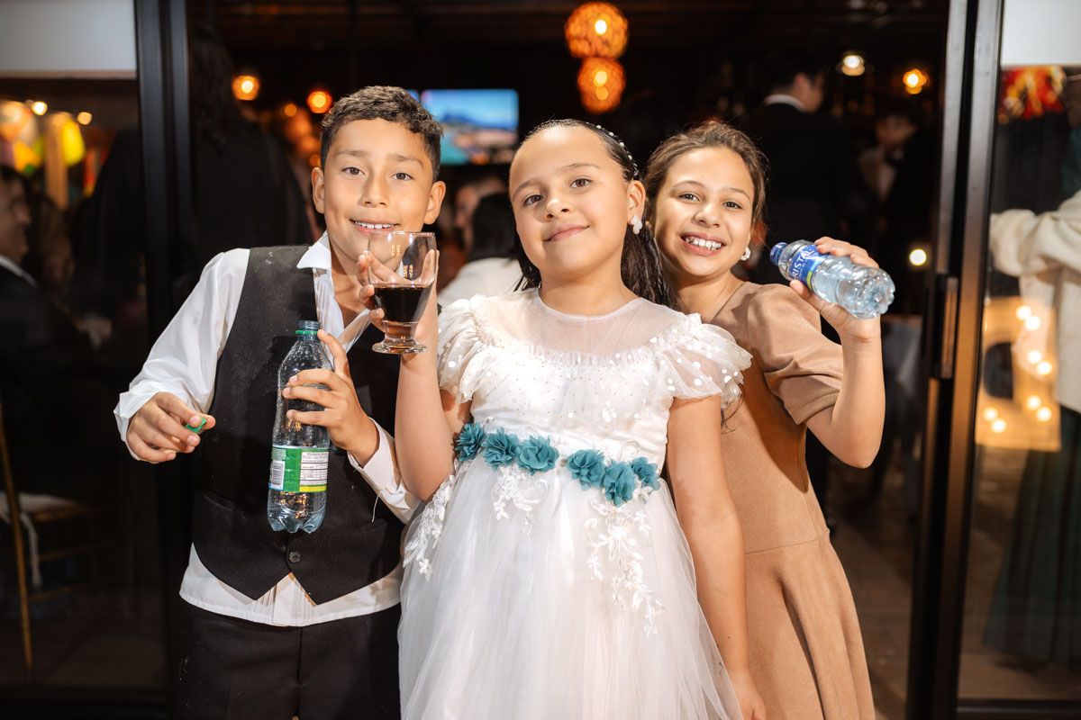 Tres niños posan sonrientes en fiesta de boda, con vestido blanco y ropa formal. Sostienen bebidas. Celebración.