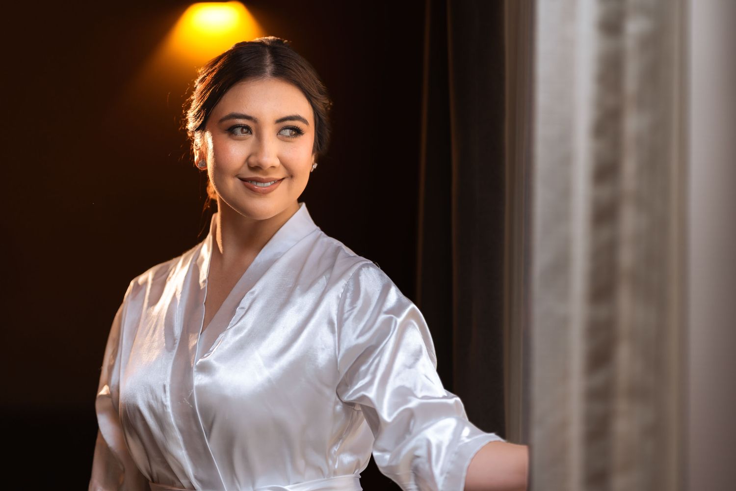 Novia sonriente con bata blanca de satín mira hacia la luz de una ventana durante su preparación de boda.