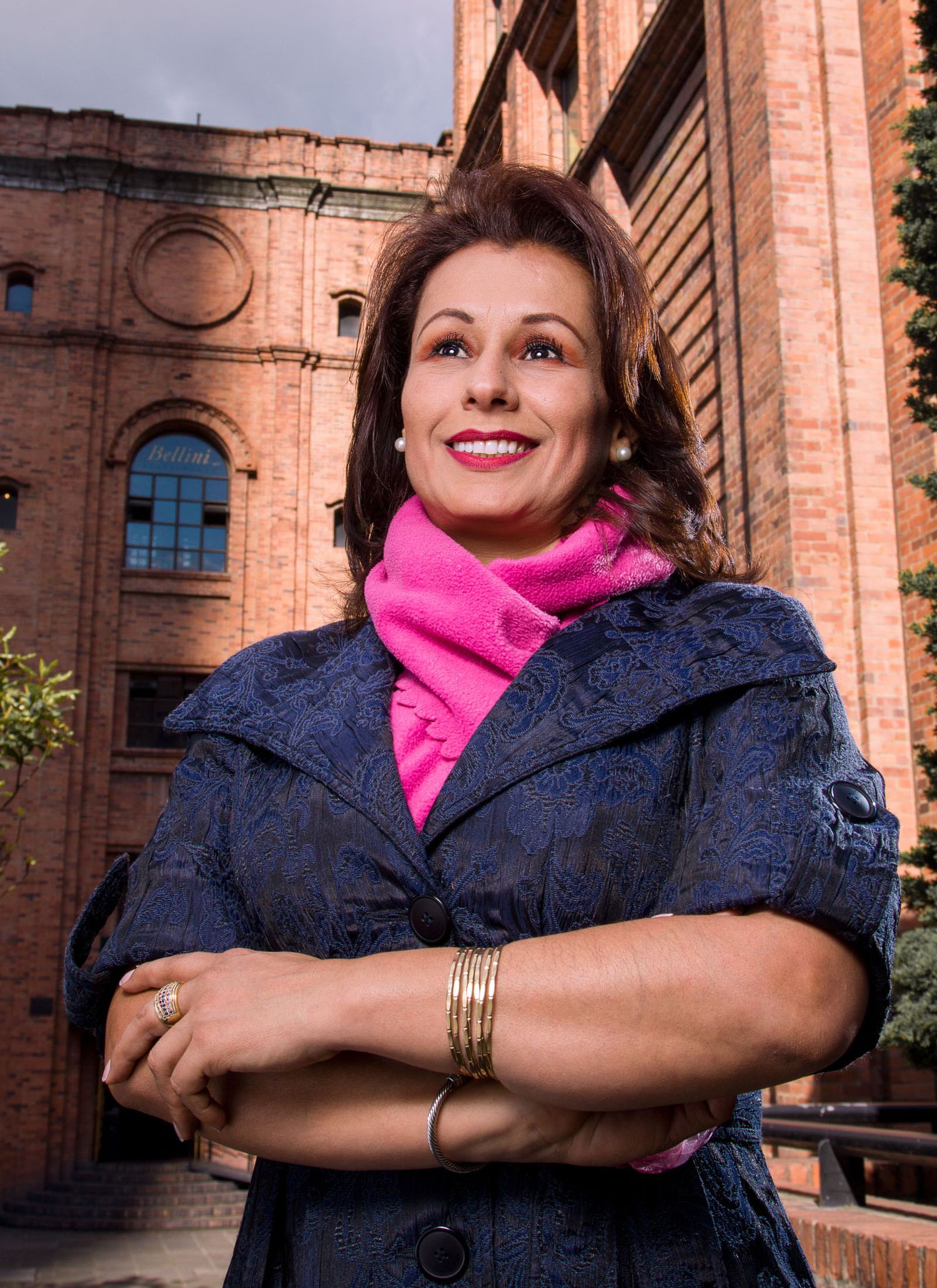 Mujer sonriente con abrigo azul y bufanda rosa posa con brazos cruzados; edificio ladrillo fondo. Retrato exterior. Olga Victoria Rubio
