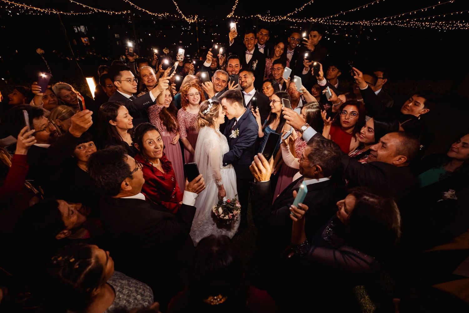 Boda nocturna romántica: recién casados besándose bajo luces cálidas, rodeados de invitados sonrientes grabando. Ambiente festivo y emotivo