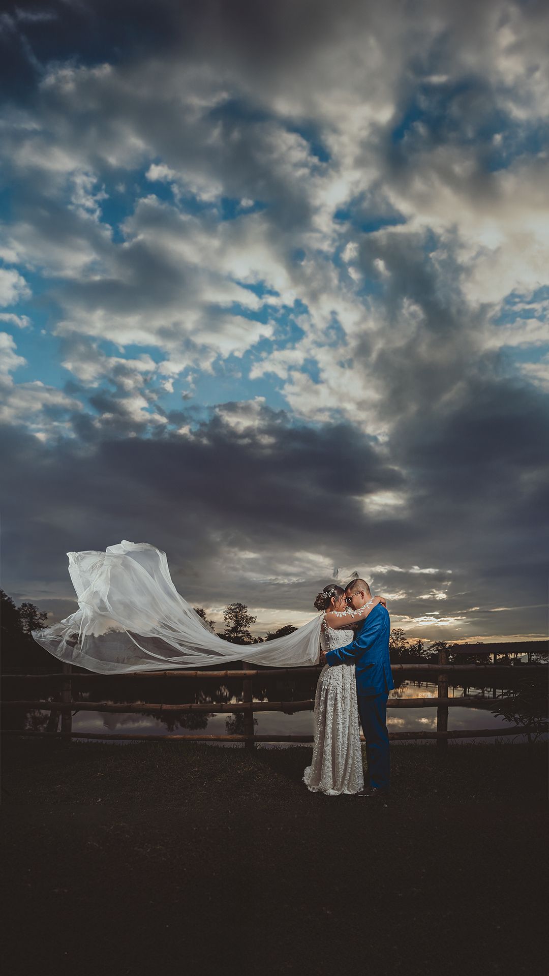 Romántica boda al atardecer: pareja abrazada en puente con velo ondeando al viento. Cielo dramático con nubes oscuras y luz dorada