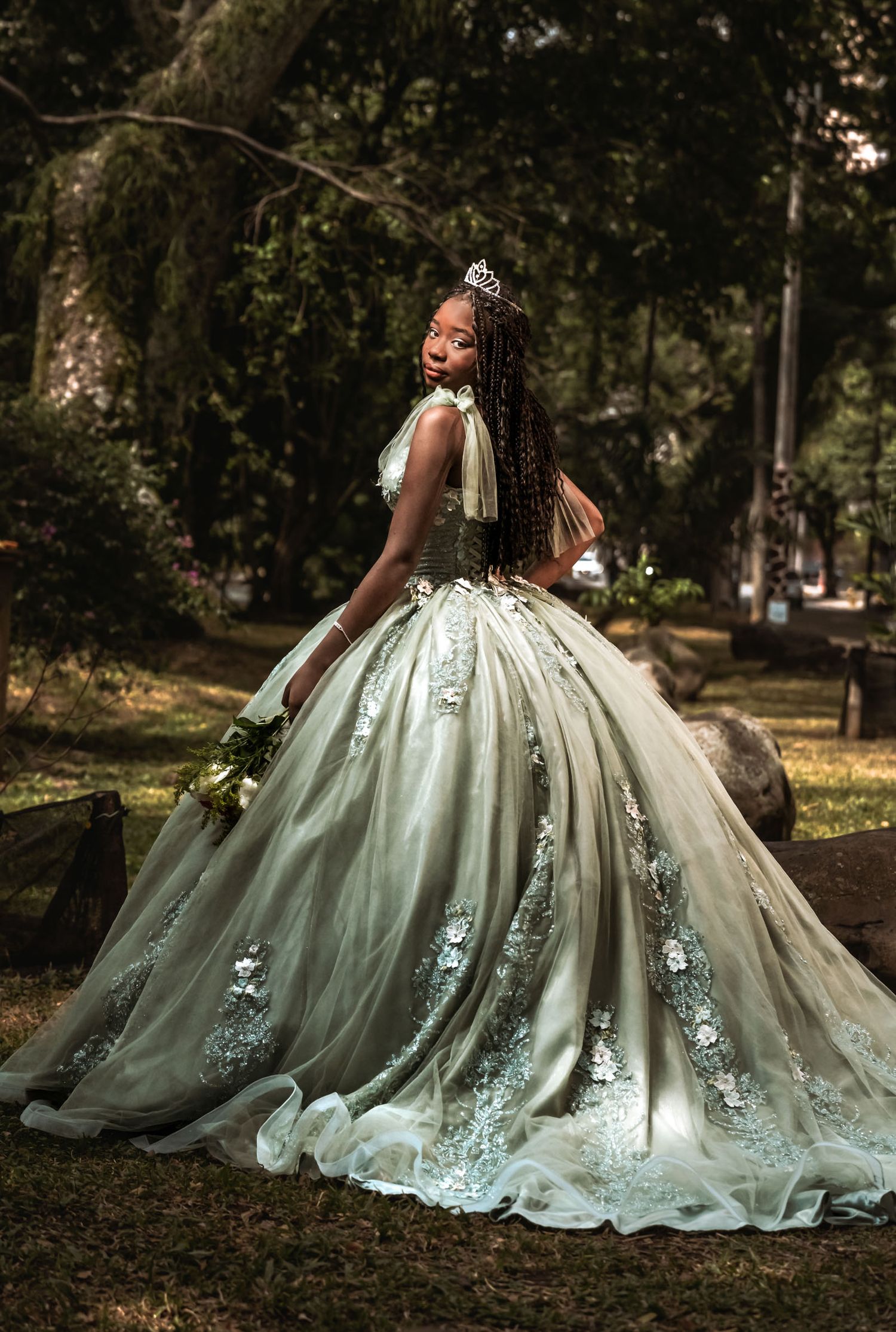 Quinceañera con vestido princesa verde/plata y tiara mira sobre hombro en parque boscoso; sesión fotos.