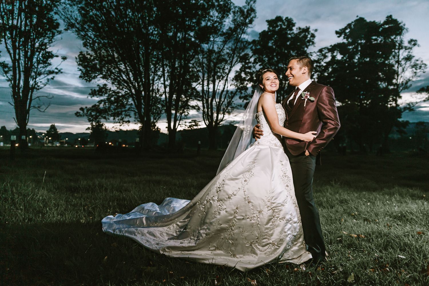 Novios se abrazan románticamente en exterior con árboles y cielo azul y nublado al anochecer. Foto de boda.