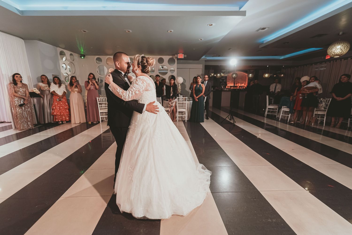 Novios comparten primer baile en pista a cuadros blanco y negro; invitados observan. Recepción de boda.