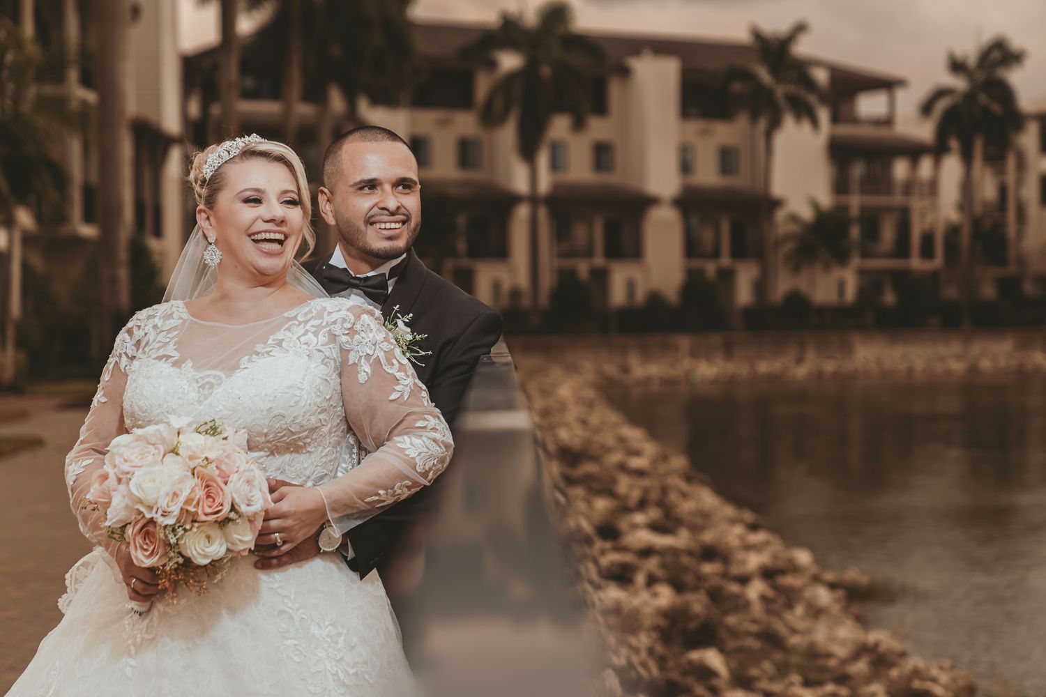 Novios posan felices junto a lago o canal; edificio residencial y palmeras al fondo. Fotografía boda en Miami.