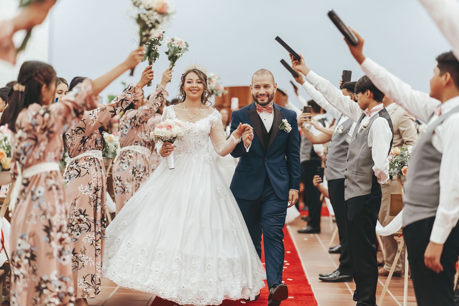Novios caminan sonrientes por pasillo con alfombra roja en la iglesia, cortejo celebra levantando ramos y brazos. Boda IDMJI en Sáchica.
