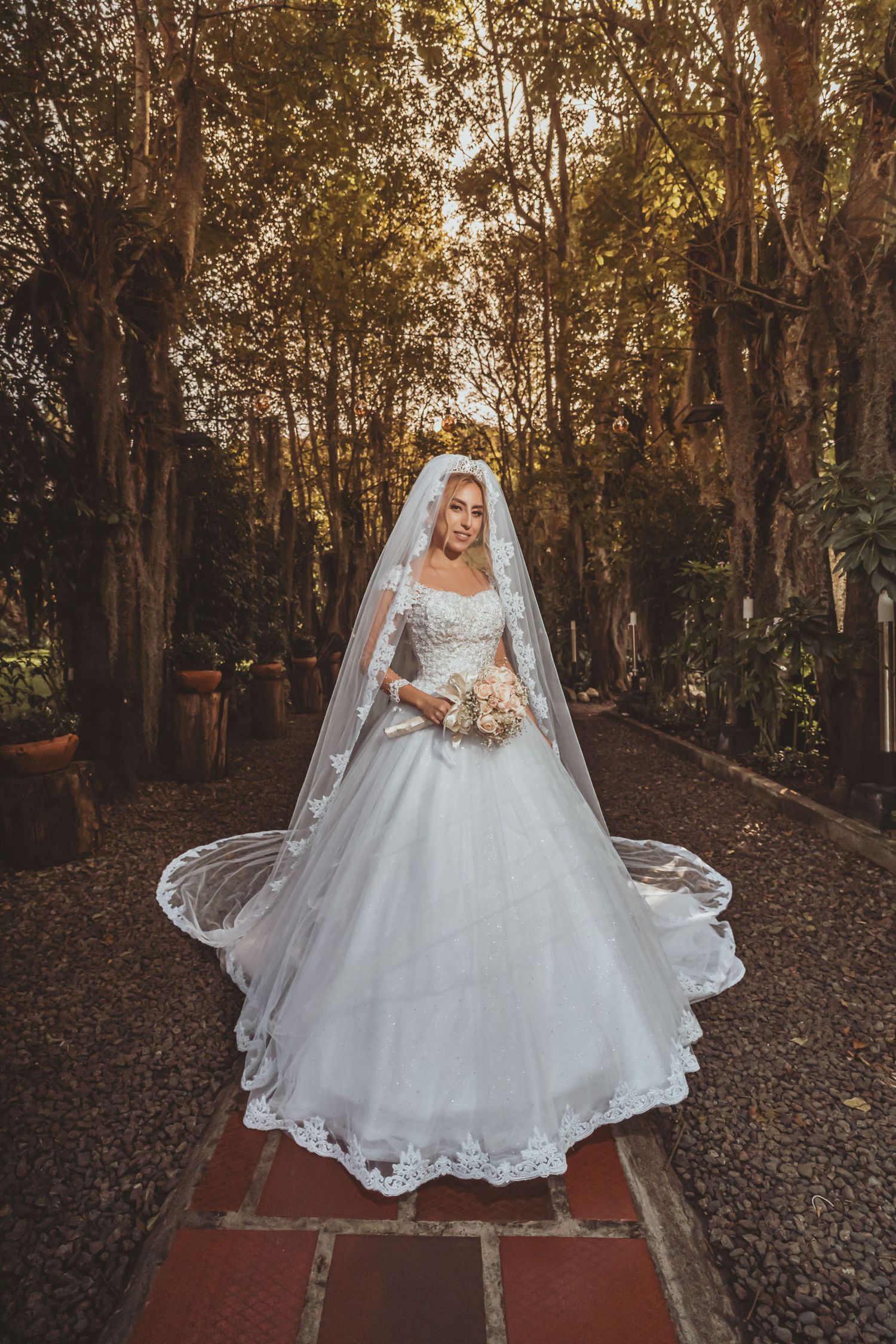 Novia con vestido princesa, velo largo y ramo posa en camino adoquinado rodeado de árboles; retrato boda.