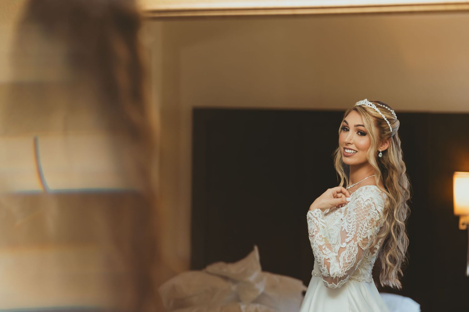 Novia sonriente con vestido de encaje y tocado mira su reflejo completo en espejo grande; preparación boda.