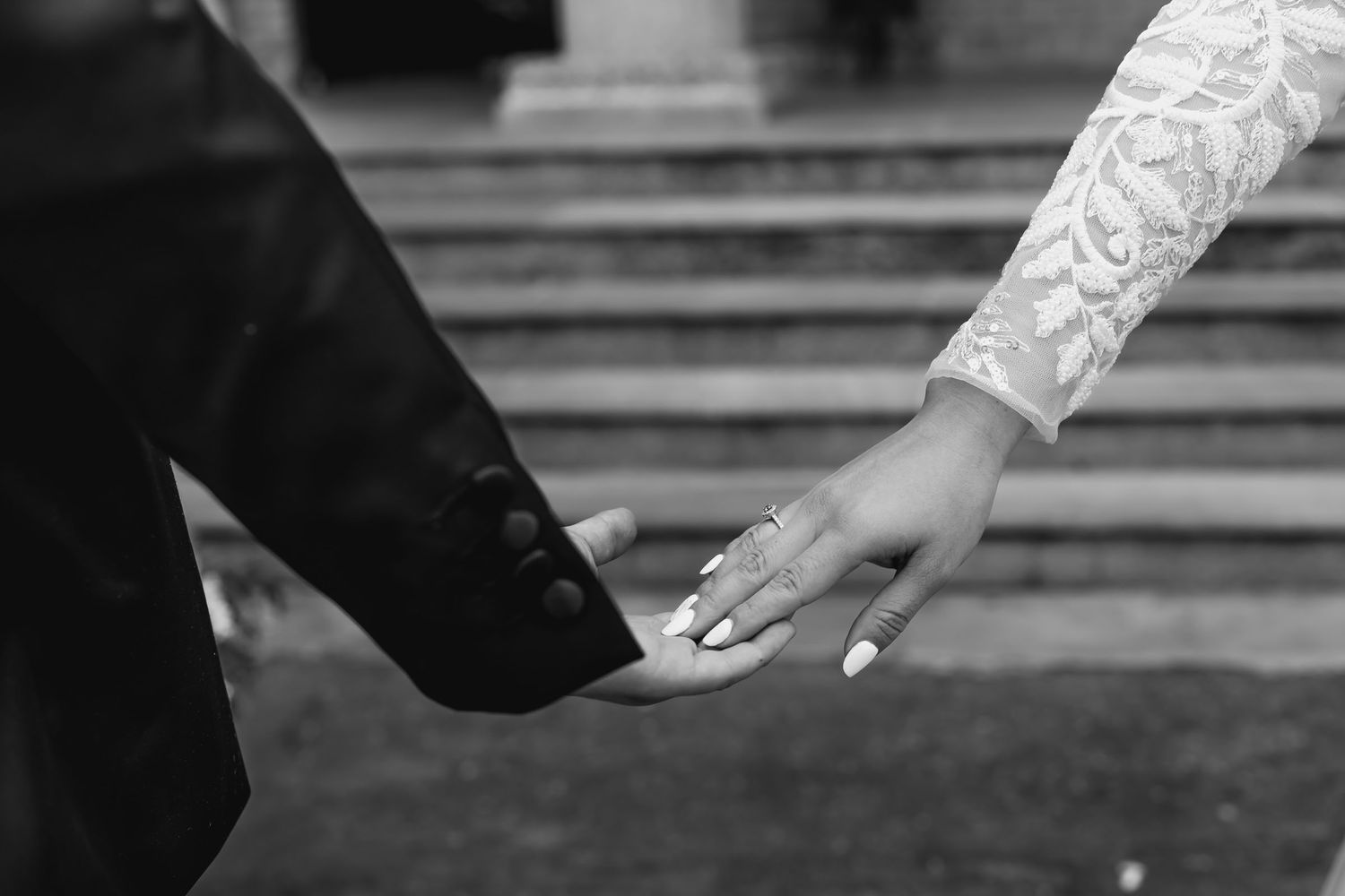 Detalle blanco y negro: manos de novios casi entrelazadas; anillo de boda visible en la mano de ella. Símbolo de unión.
