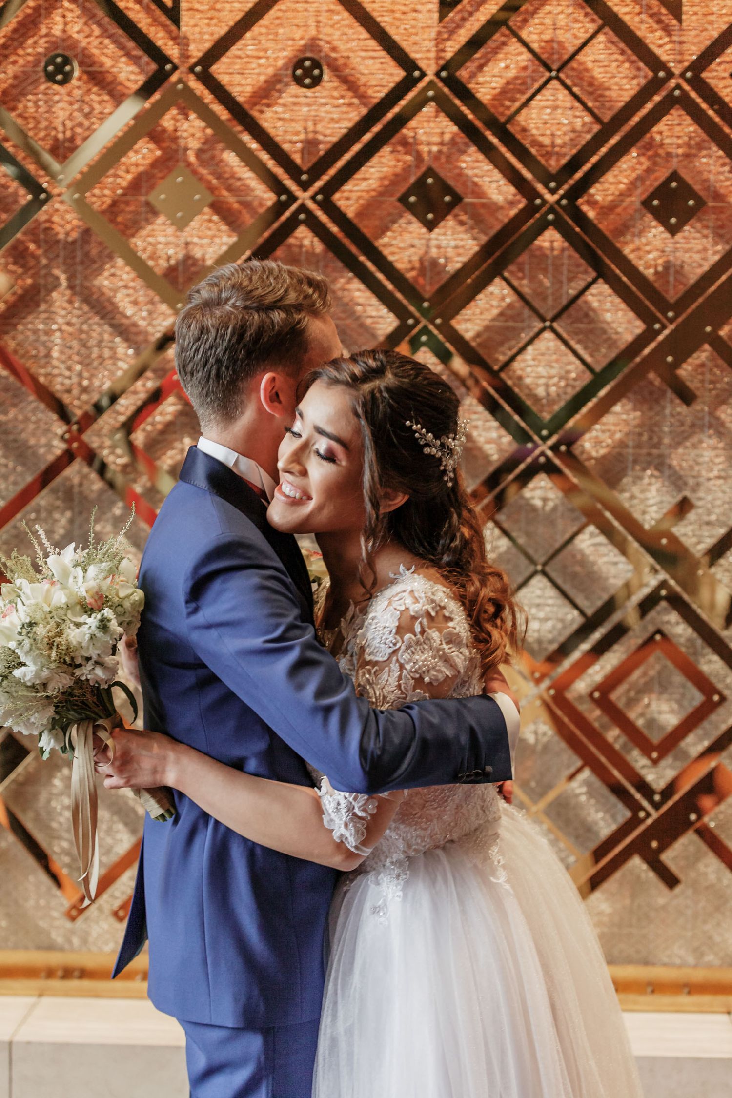Novios se abrazan felices y con ternura tras 'first look'; panel dorado fondo. Novia sostiene ramo.
