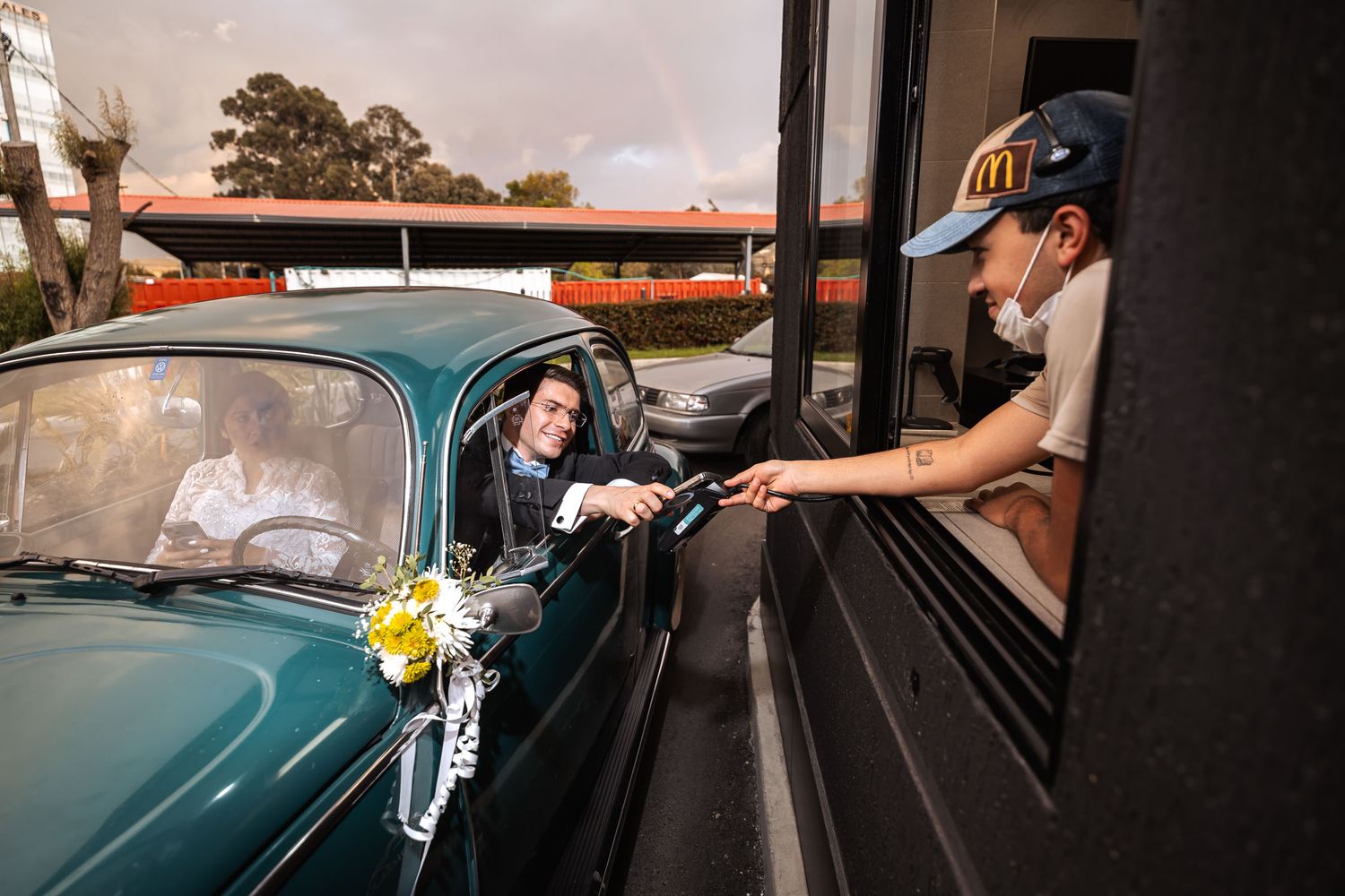 Novios en coche clásico verde reciben algo en ventanilla de servicio al volante. Momento divertido y único en su boda.