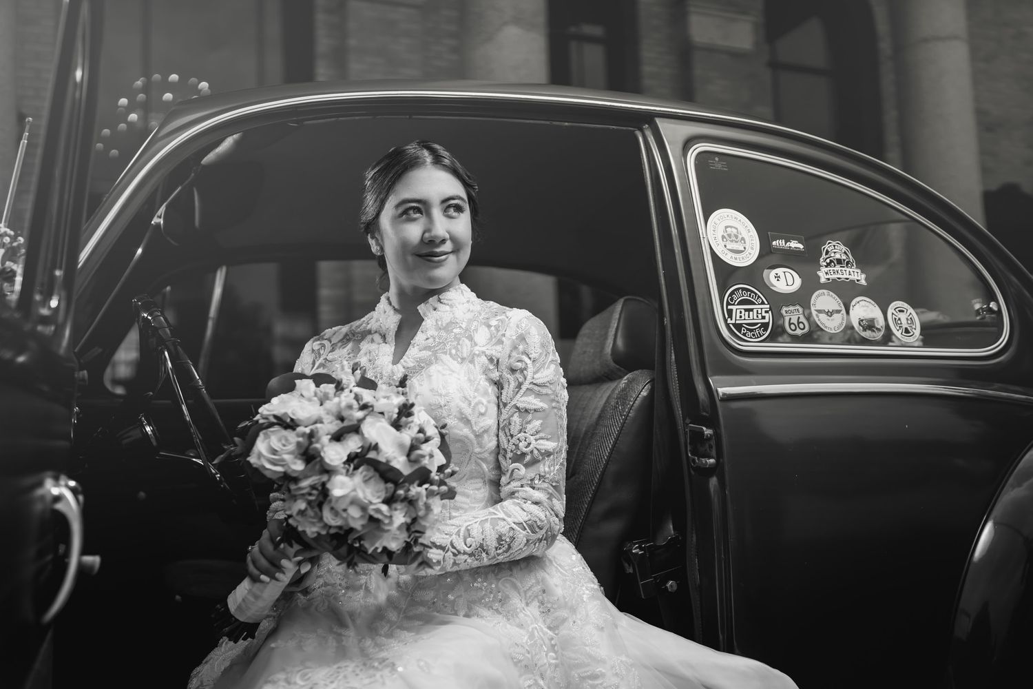 Foto blanco y negro: Novia sonriente con vestido encaje y ramo sentada en puerta de coche clásico VW Escarabajo. Boda.