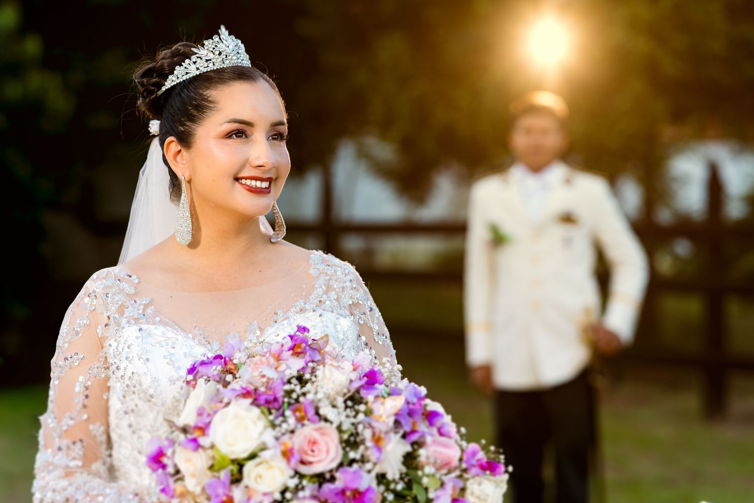 Retrato de la novia con su ramo y tiara, con el novio desenfocado en el fondo al atardecer