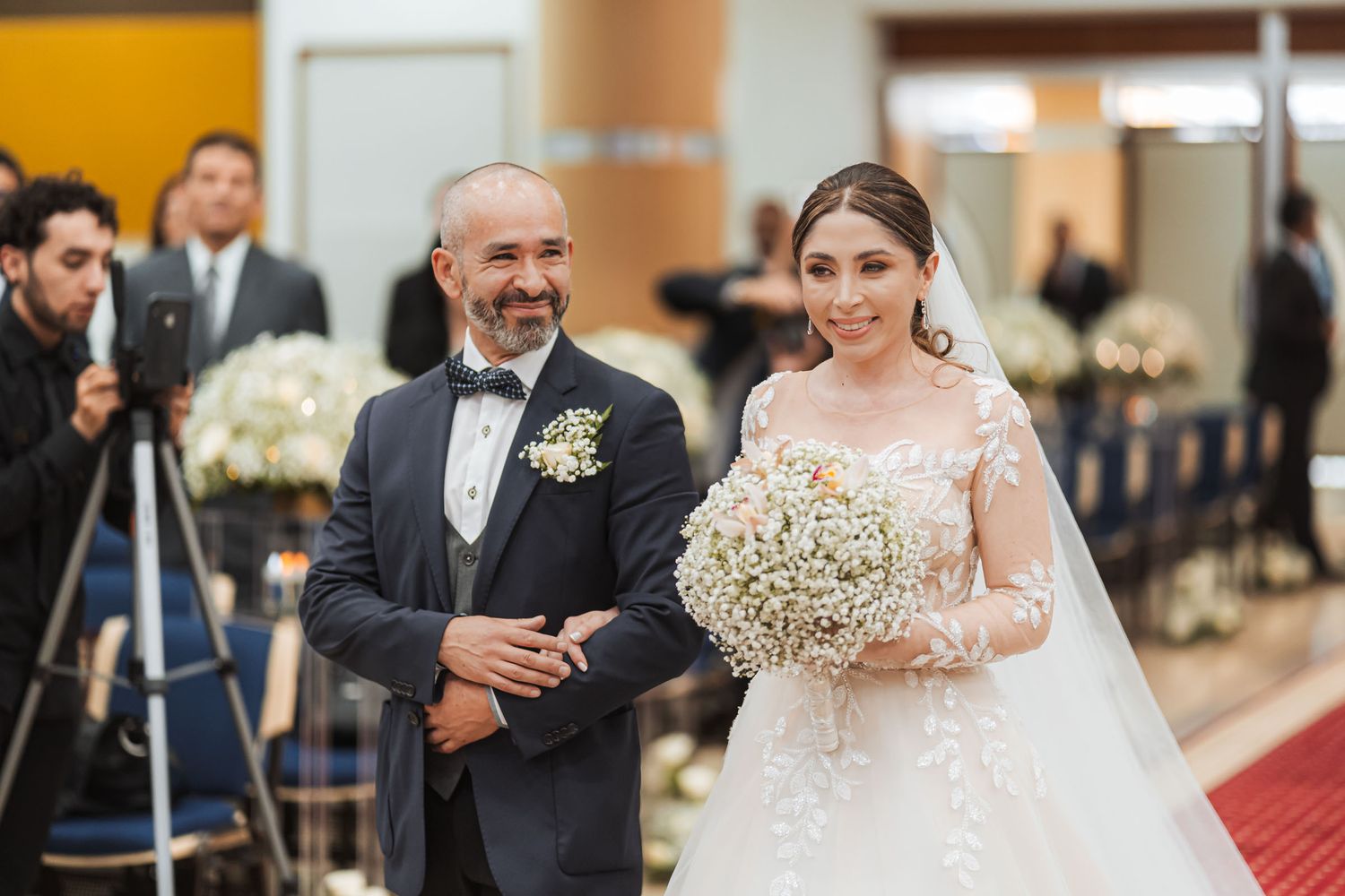 Novia sonriente camina al altar del brazo de su padre (o padrino) durante entrada nupcial en iglesia. Boda IDMJI.