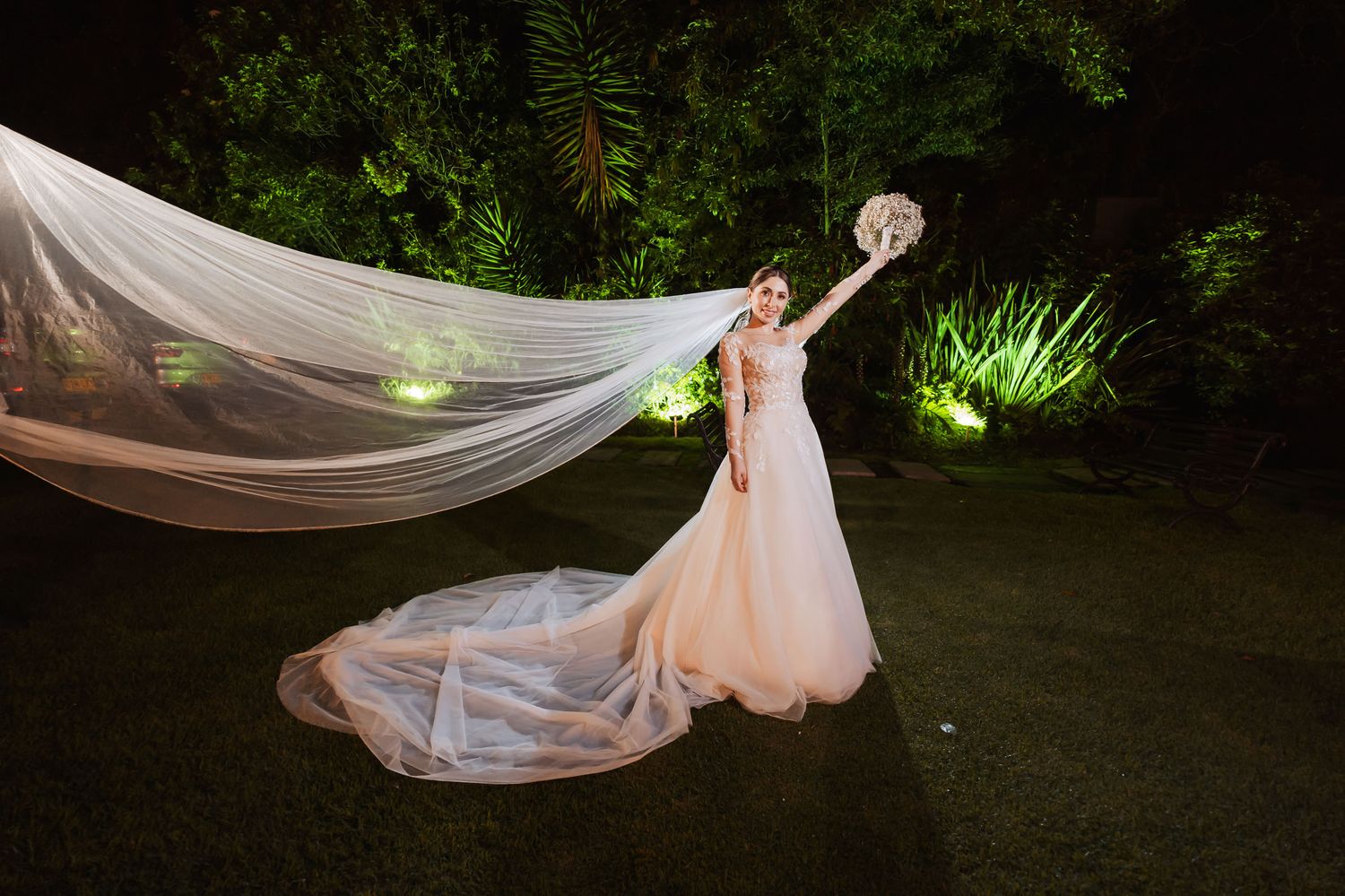 Novia sola de noche en jardín lanza su ramo al aire; velo largo flota espectacularmente. Foto boda creativa.
