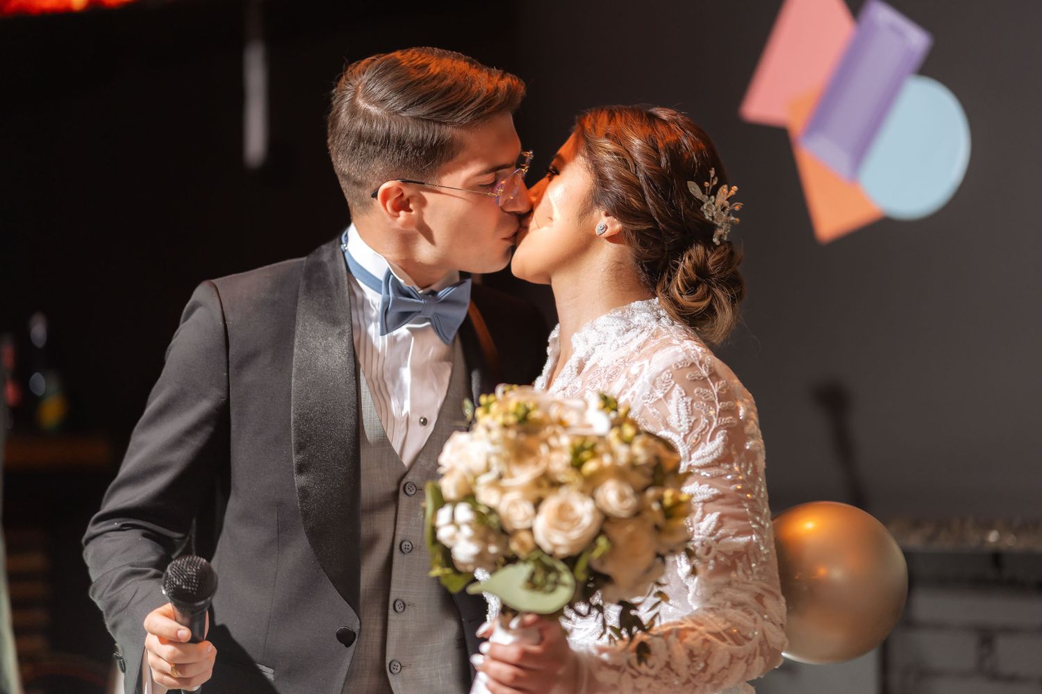 Novios se besan en escenario durante la recepción de boda, sosteniendo micrófono y ramo. Decoración colorida.