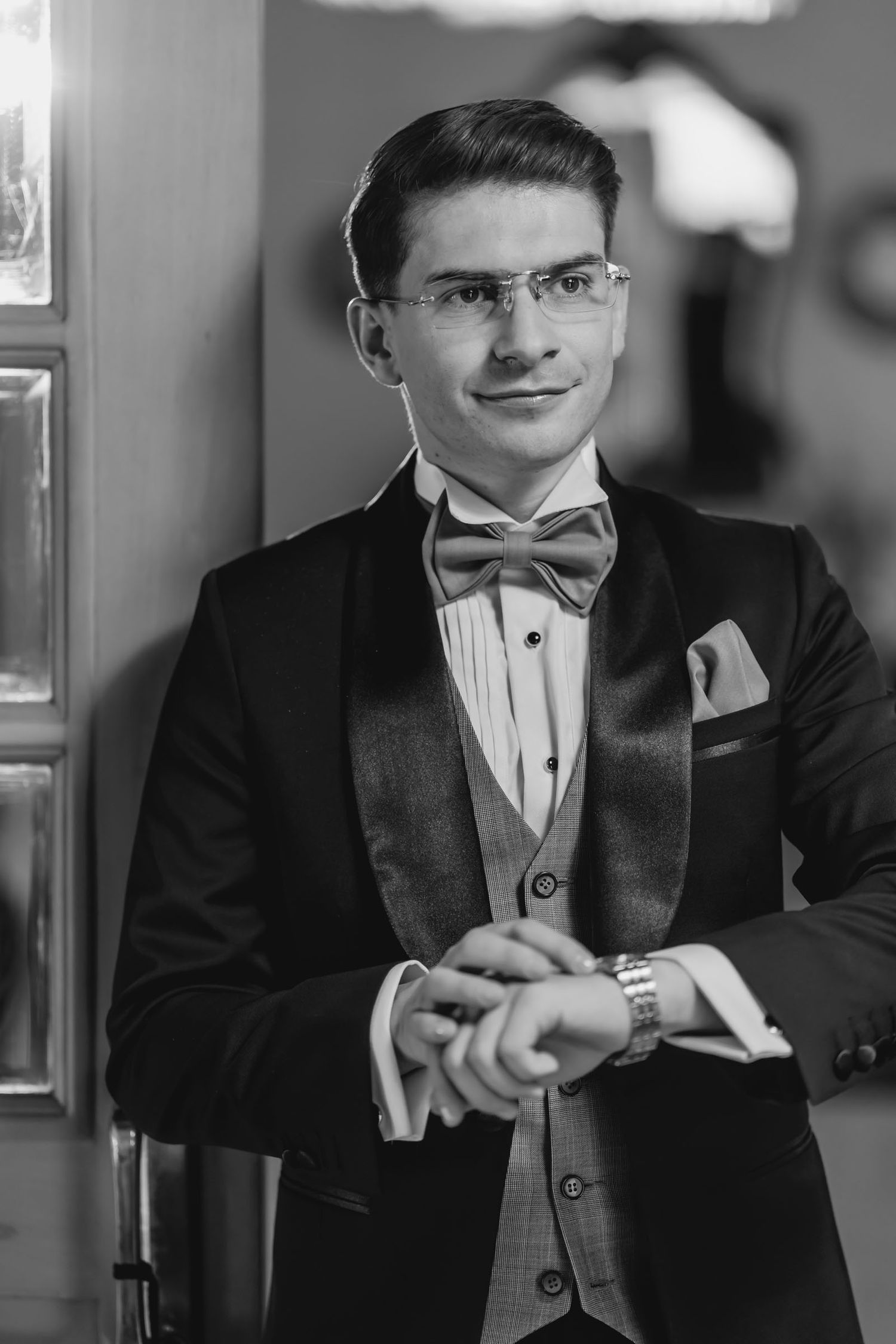 Foto blanco y negro: Novio con esmoquin ajusta su reloj; retrato elegante durante preparación de boda.