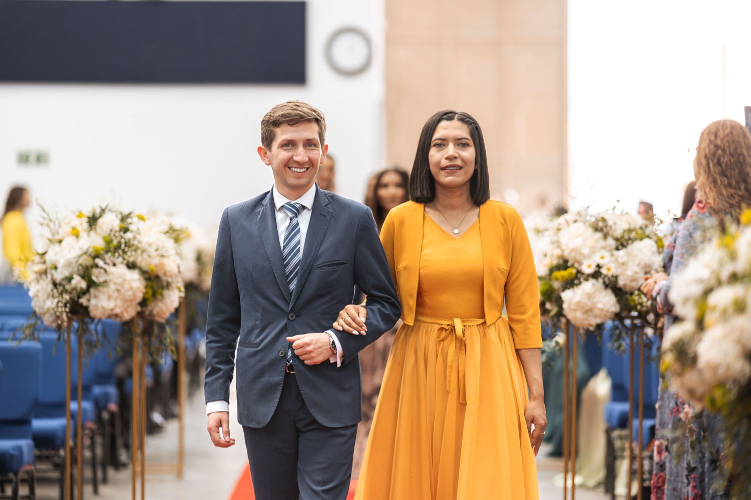 Hombre con traje azul camina del brazo con mujer de vestido amarillo mostaza por pasillo de iglesia; procesión de boda.