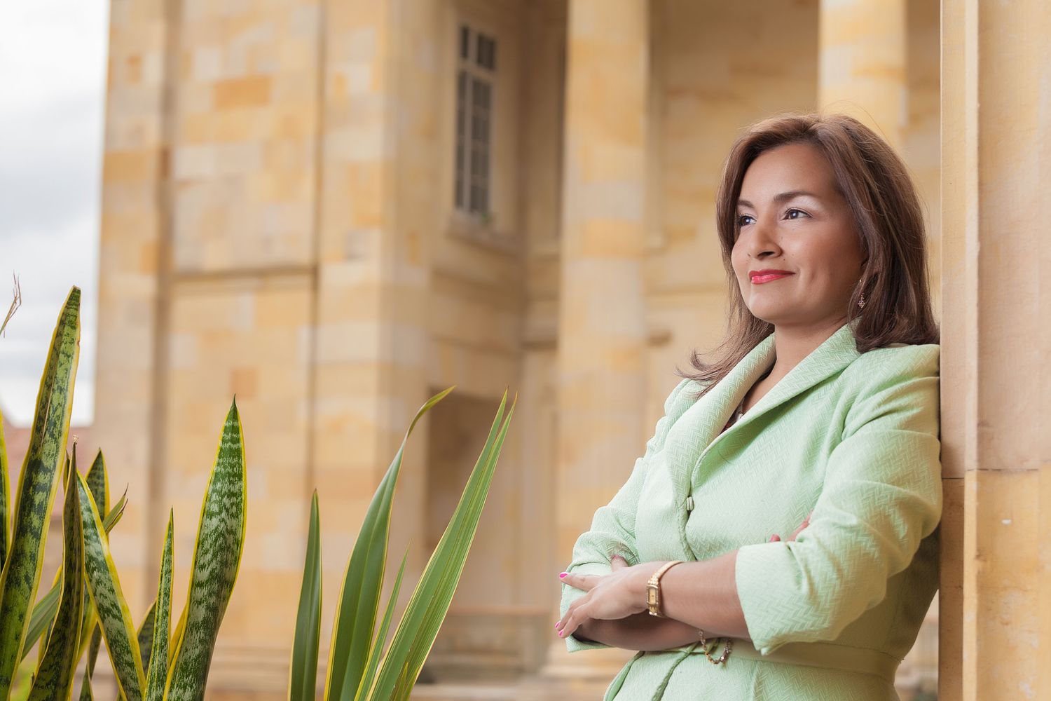 Mujer congresista sonríe con brazos cruzados, vistiendo blazer verde, frente a edificio del congreso y arquitectura clásica con columnas.