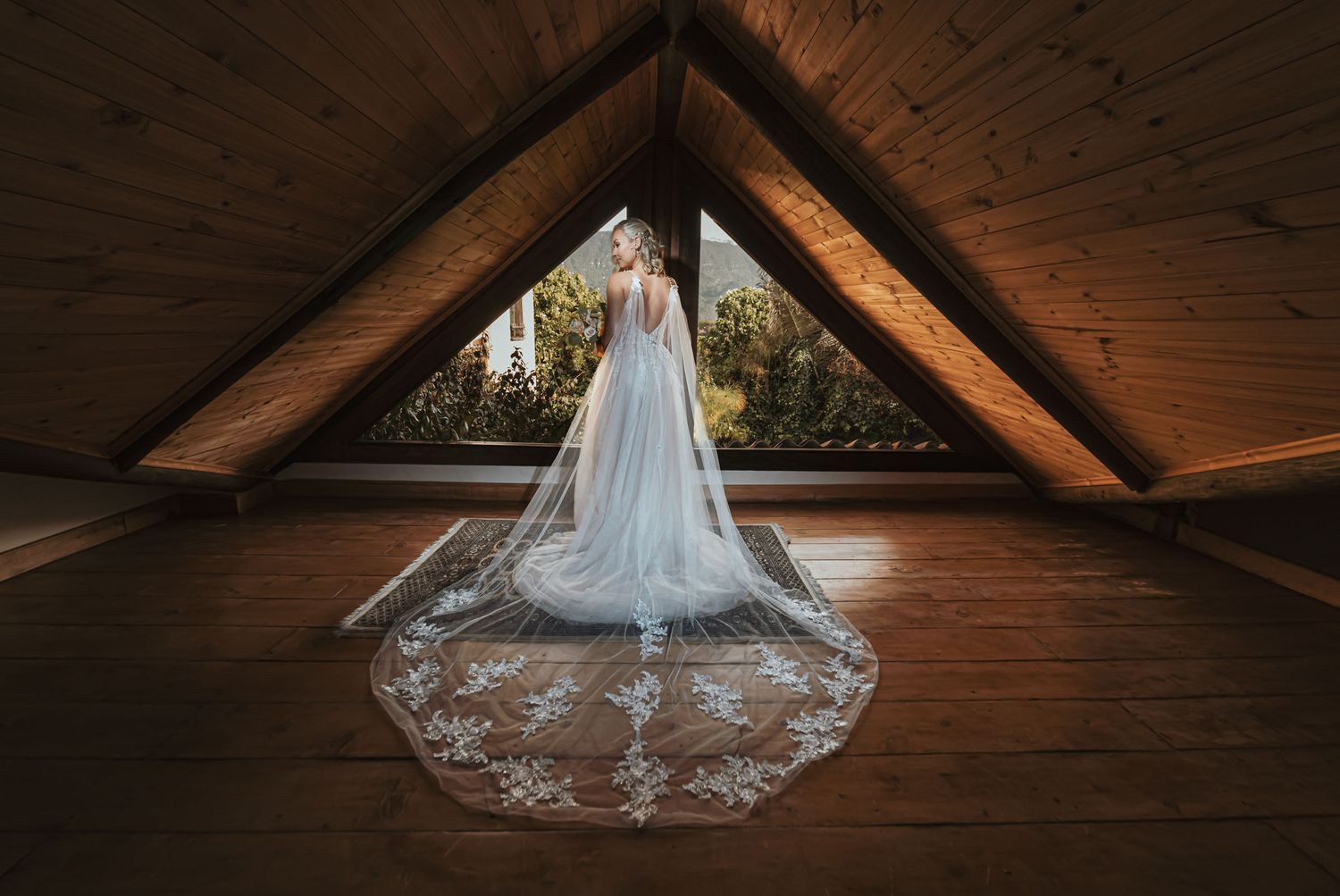 Novia solitaria de espaldas en ático rústico con luz natural entrando por ventana triangular. Viste elegante vestido blanco con velo bordado