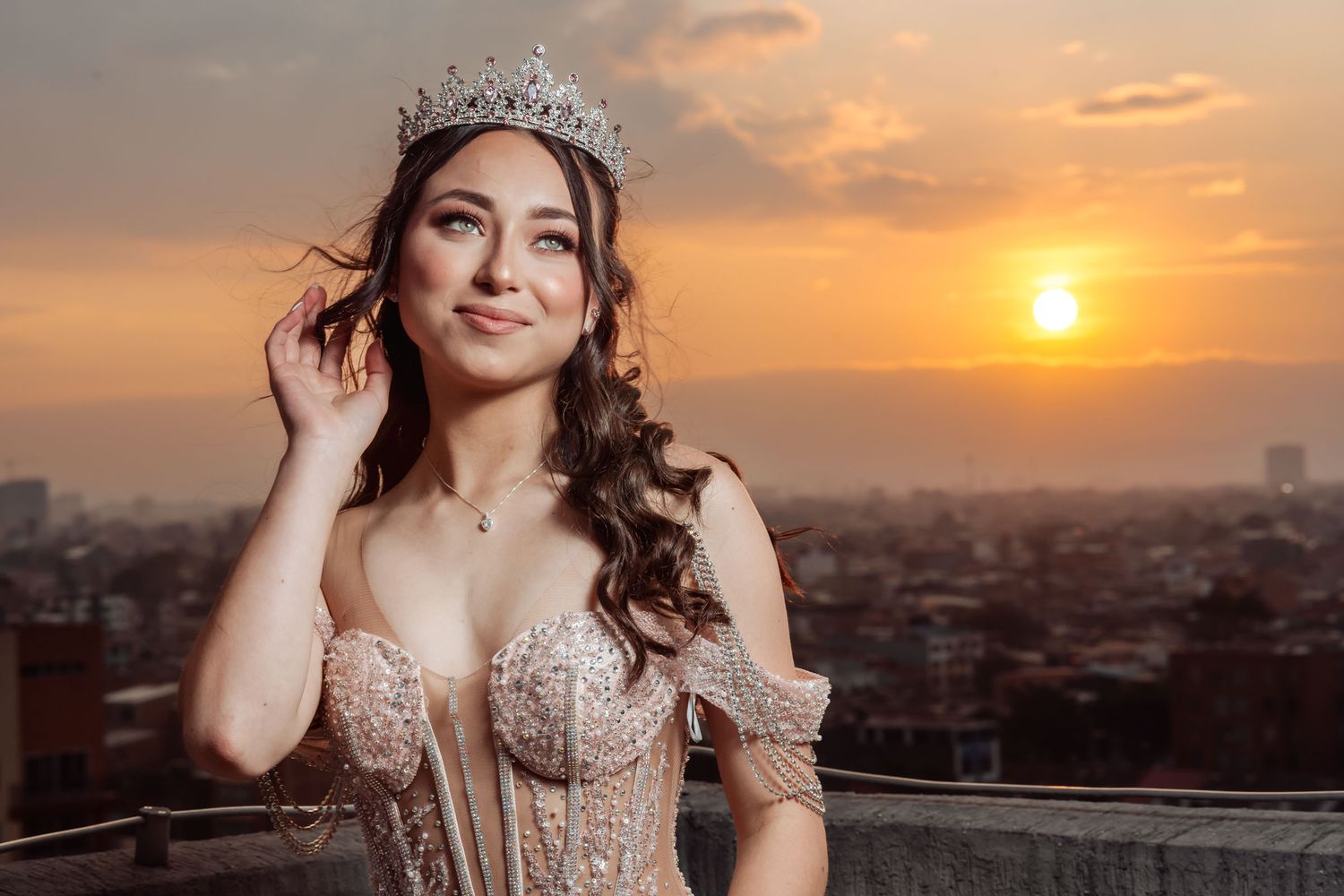 Retrato quinceañera sonriente (vestido corsé rosa, tiara) tocando cabello en terraza al atardecer sobre ciudad y atardecer naranja de fondo