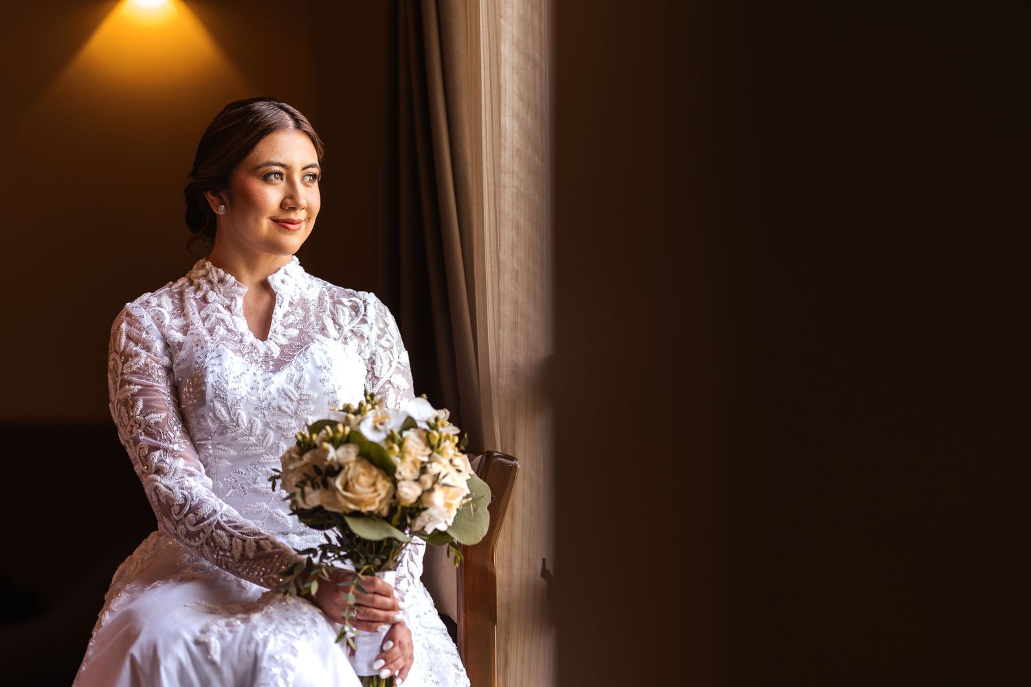 Novia con vestido de novia de encaje sostiene su ramo sentada junto a ventana; retrato con luz natural antes de la boda.