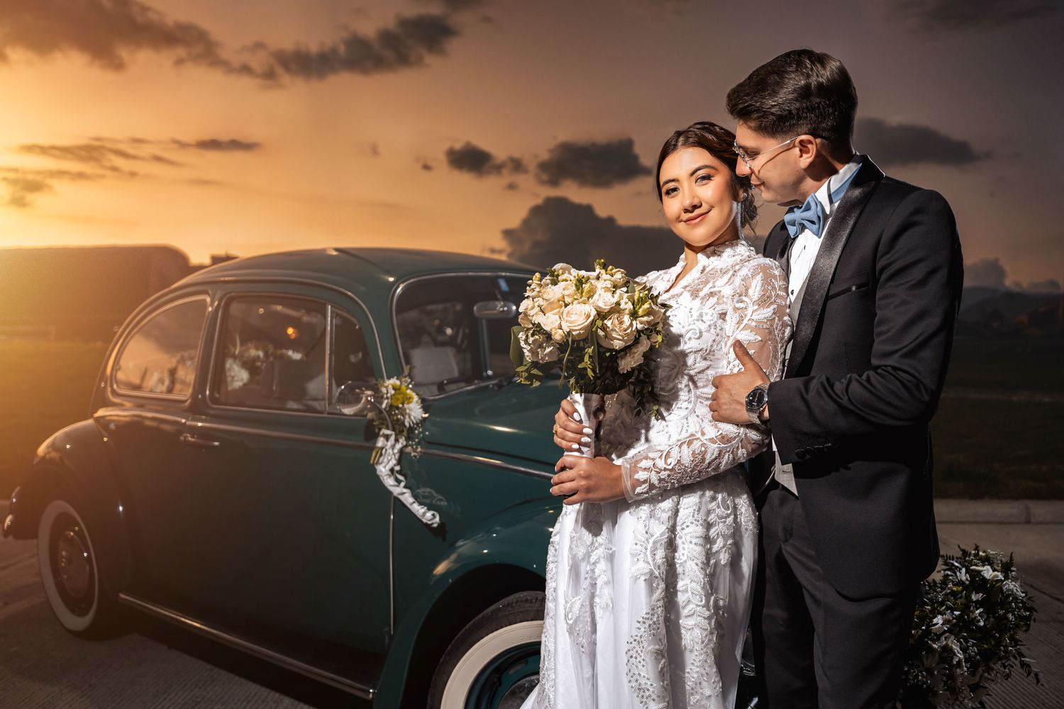 Novios tomados de la mano miran paisaje al atardecer junto a coche verde clásico. Abrazo tierno en boda.