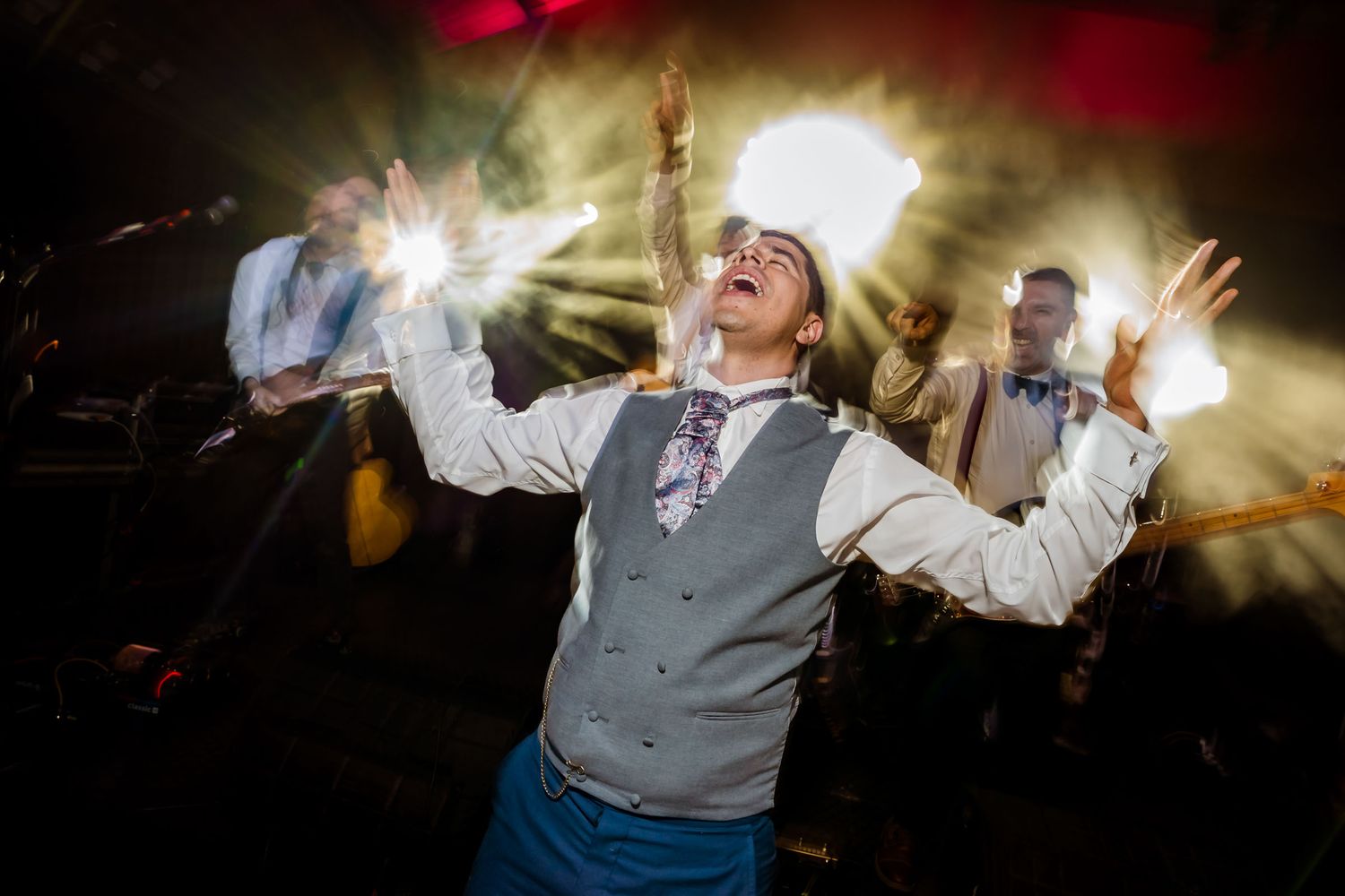 Enérgico novio celebra en la recepción, brazos alzados y ojos cerrados cantando bajo focos brillantes. Música en vivo.