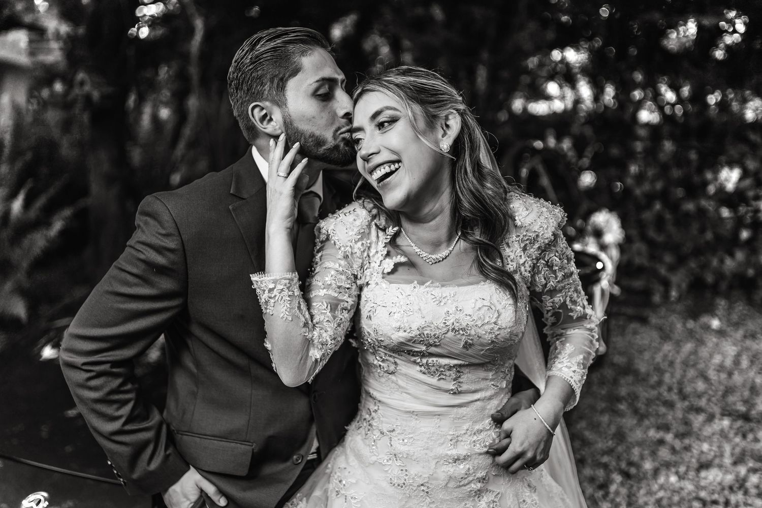 Foto B&W boda: Novio besa tiernamente sien de novia sonriente; retrato íntimo exterior jardín.