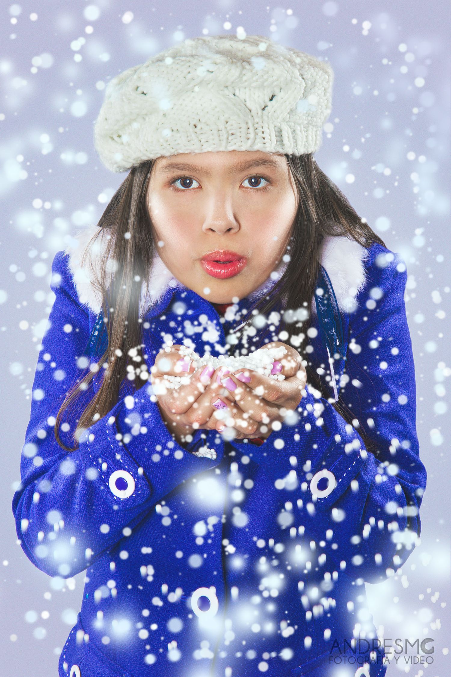 Joven con abrigo azul y boina blanca sopla nieve artificial/brillo de sus manos; retrato estudio invierno.