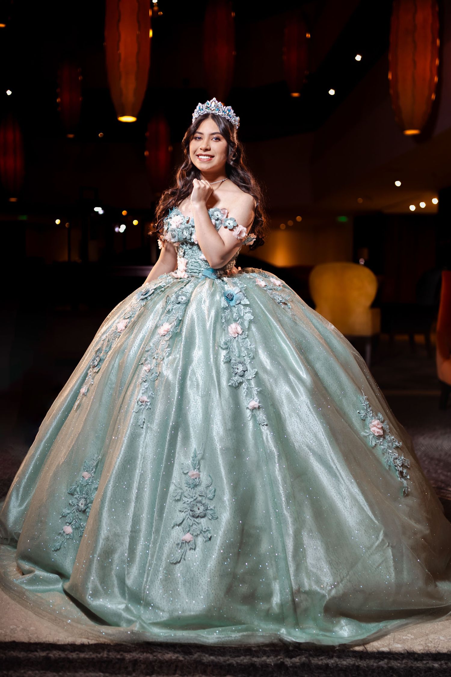 Quinceañera sonriente con voluminoso vestido princesa azul (bordado flores) y tiara; salón con luces cálidas.