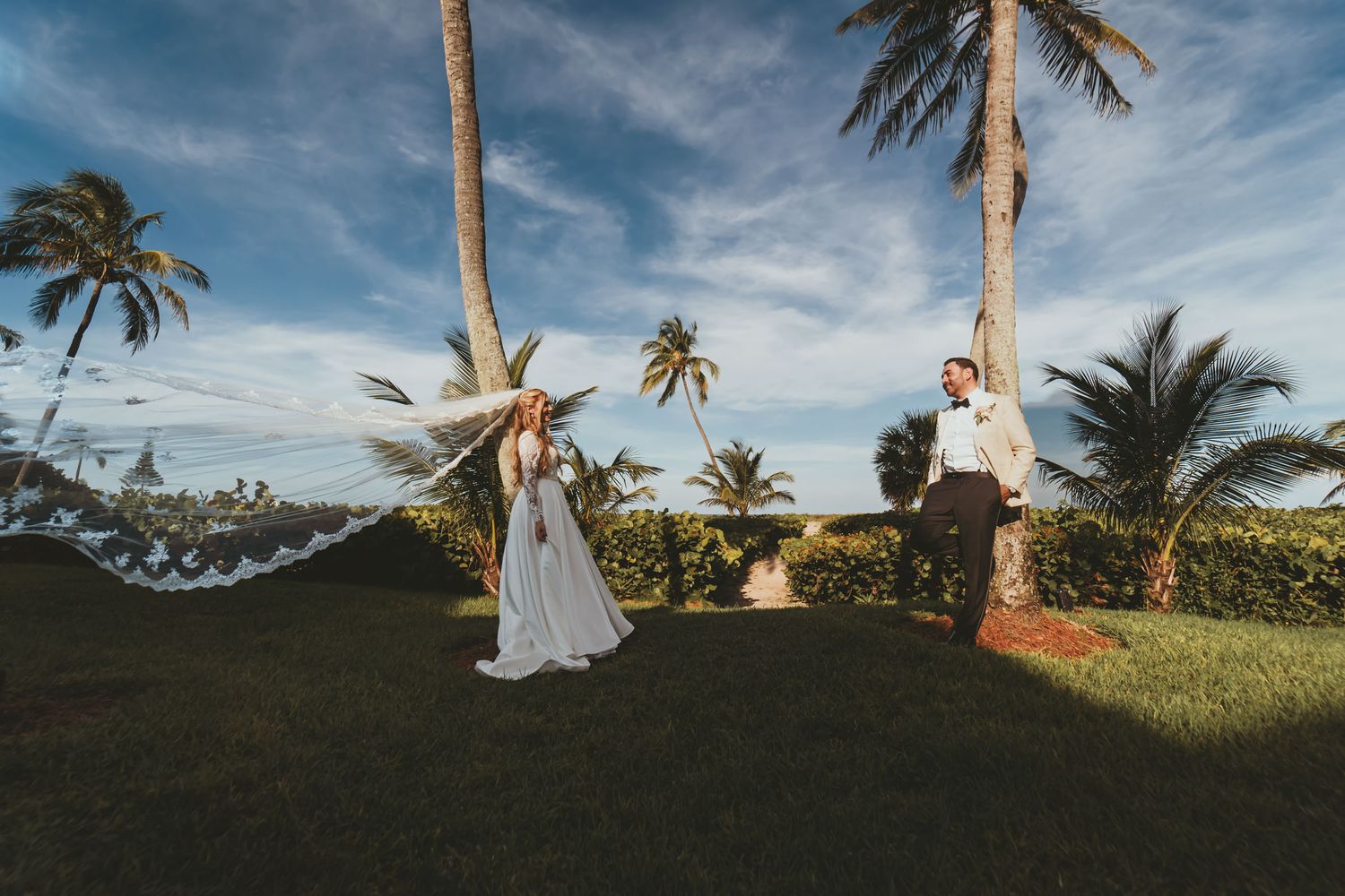 Novia con velo largo al viento y novio posan en césped bajo palmeras; cielo azul. Boda tropical.