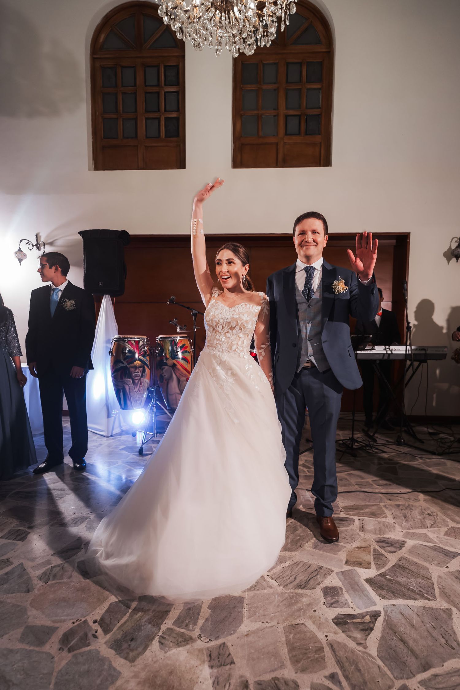 Novia y novio saludan felices con brazos en alto en pista de baile durante recepción de boda; ella vestido princesa.