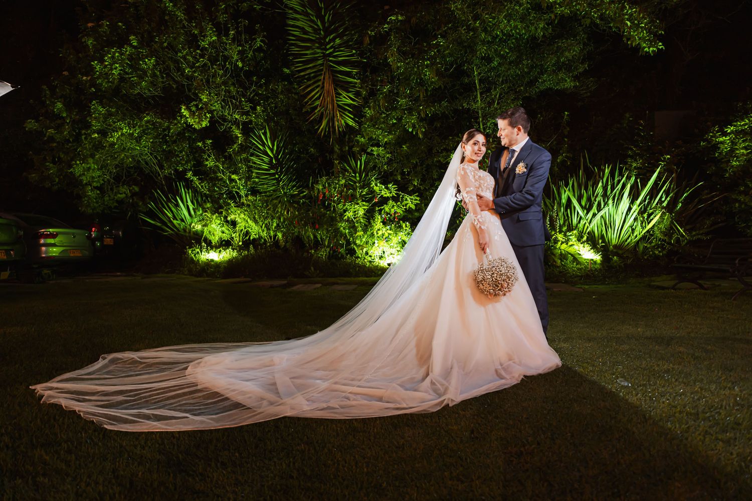 Novios posan de noche en jardín iluminado; largo velo de novia extendido sobre el césped. Fotografía de boda.