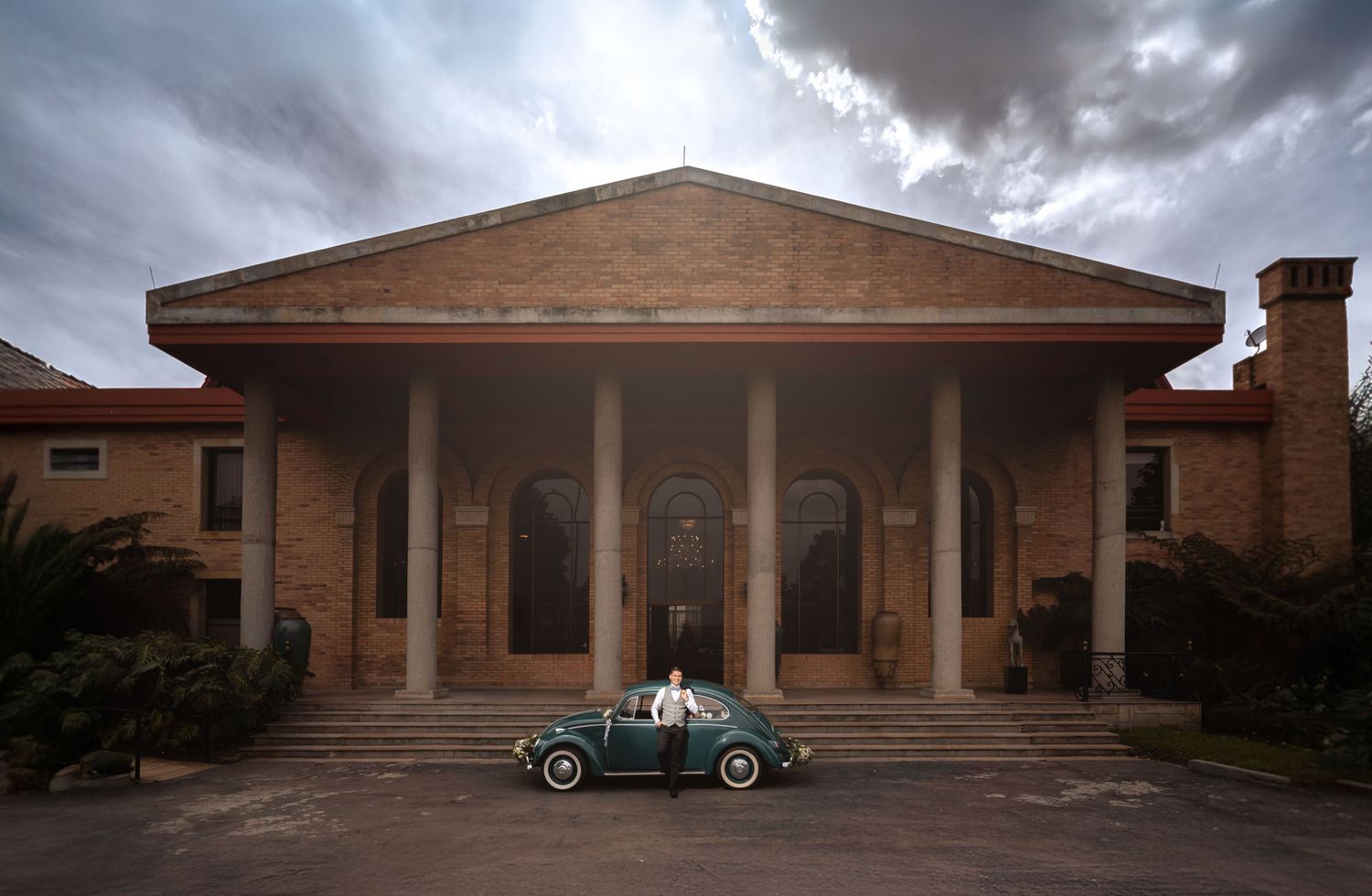 Novio con esmoquin posa junto a VW Escarabajo verde frente a gran edificio con columnas bajo cielo nublado; foto boda.