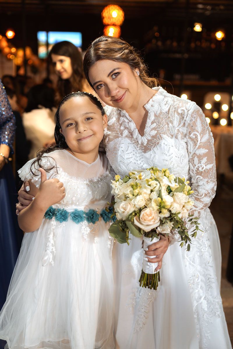 Novia sonríe posando con niña en vestido blanco y azul en su boda. Sostiene ramo. Momento tierno.