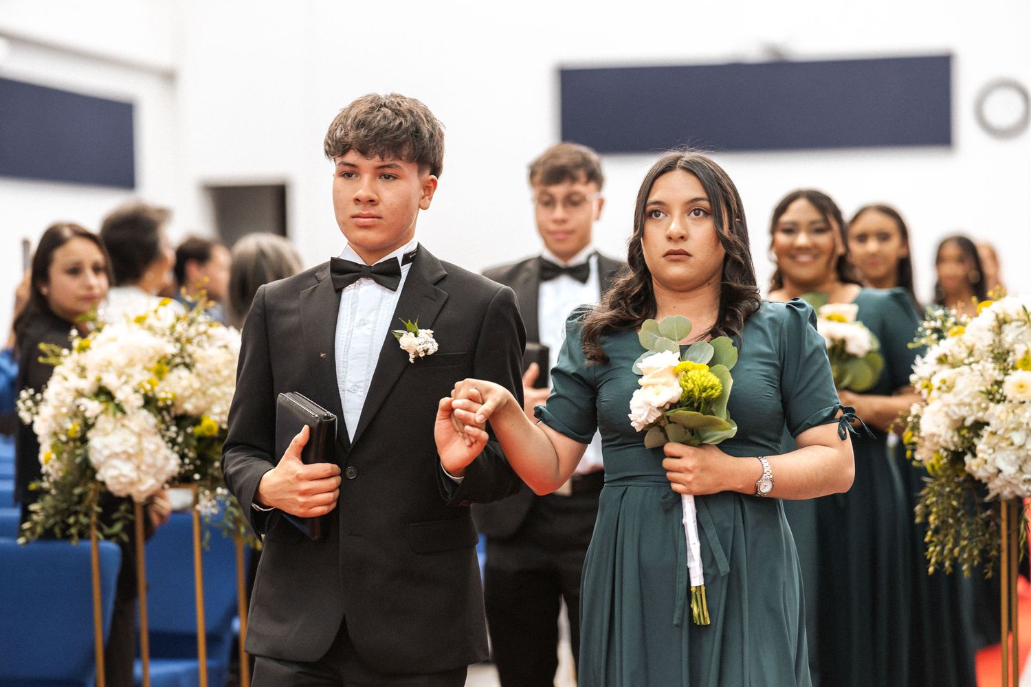 Dama de honor en vestido verde y caballero caminan por pasillo en ceremonia de boda IDMJI, tomados de la mano.