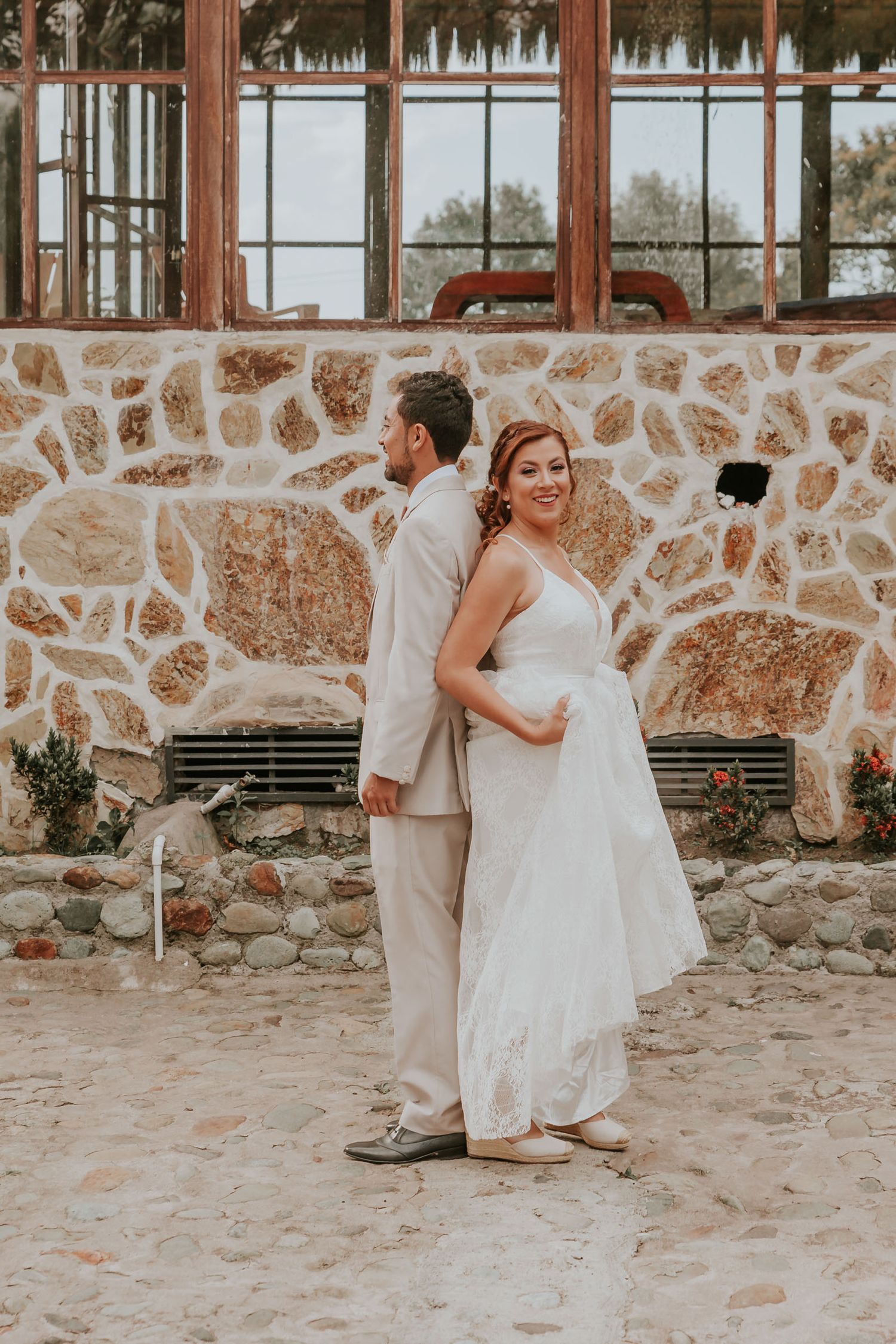 Novios posan espalda con espalda antes de 'first look'; él traje claro, ella vestido blanco. Muro piedra.