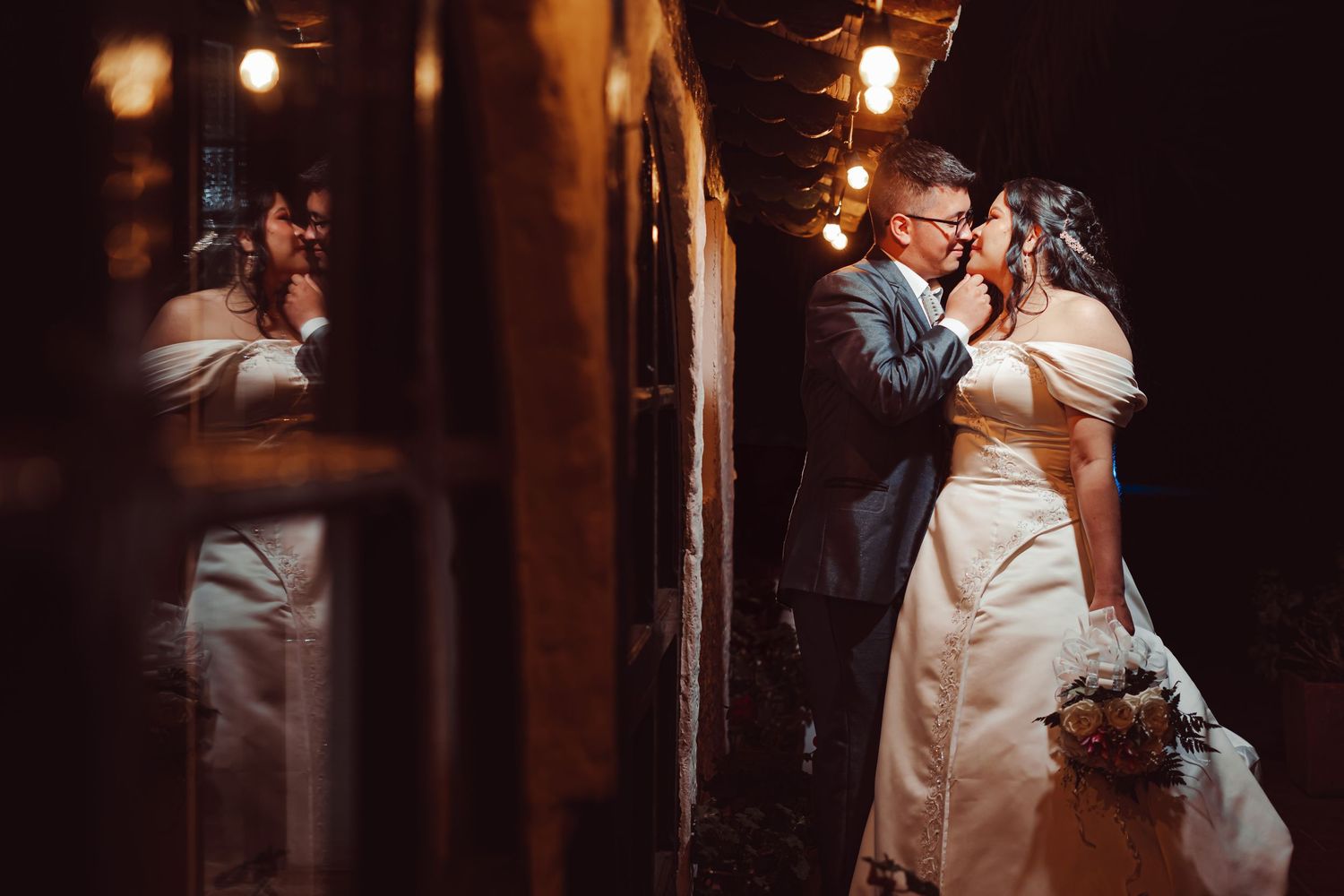 Novios casi besándose reflejados en ventana de noche; luz cálida interior. Momento íntimo boda.