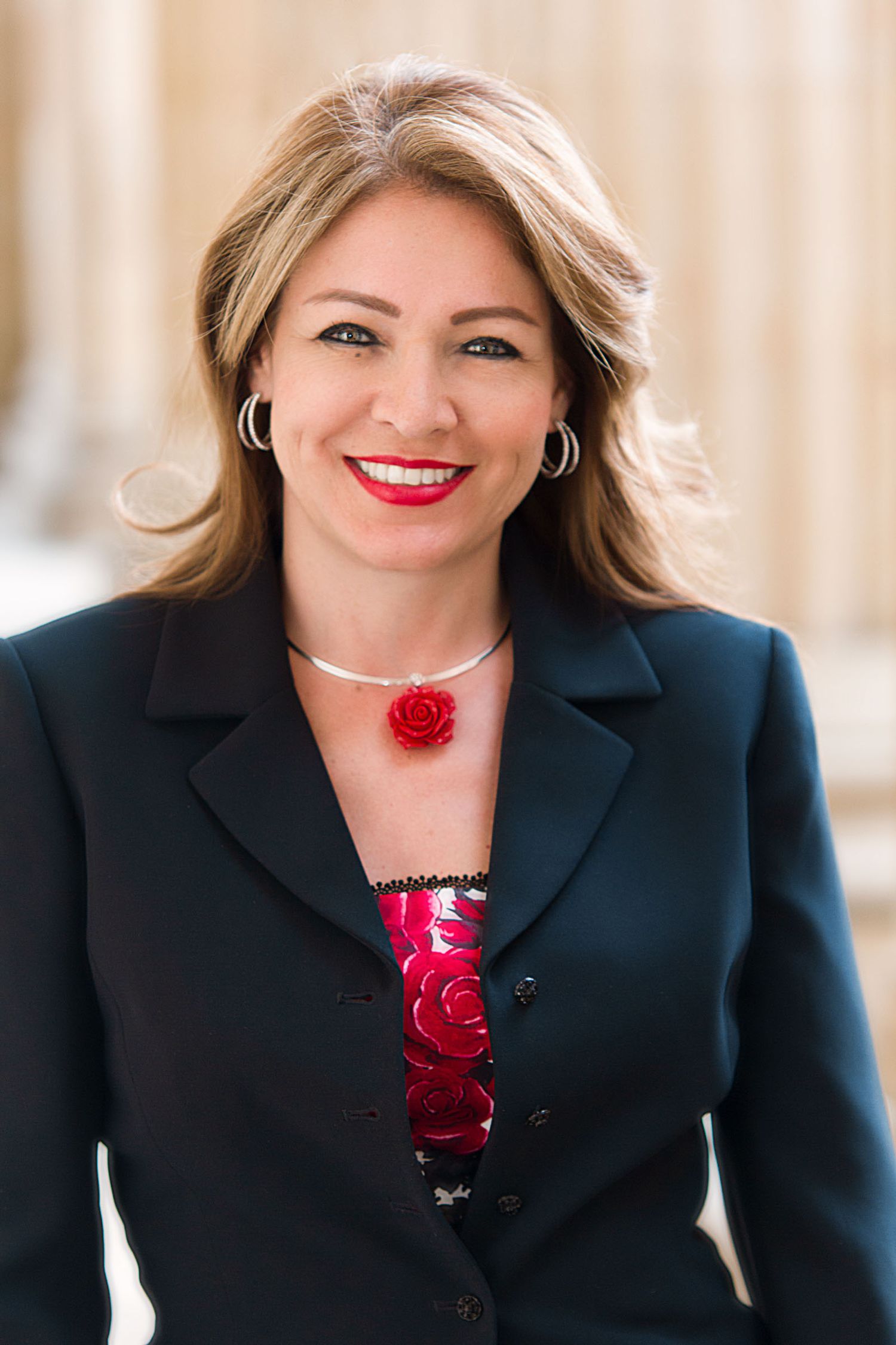 Retrato mujer profesional sonriente; lleva blazer negro sobre top rojo, collar rojo. Fondo exterior columnas. Alexandra Moreno Piraquive