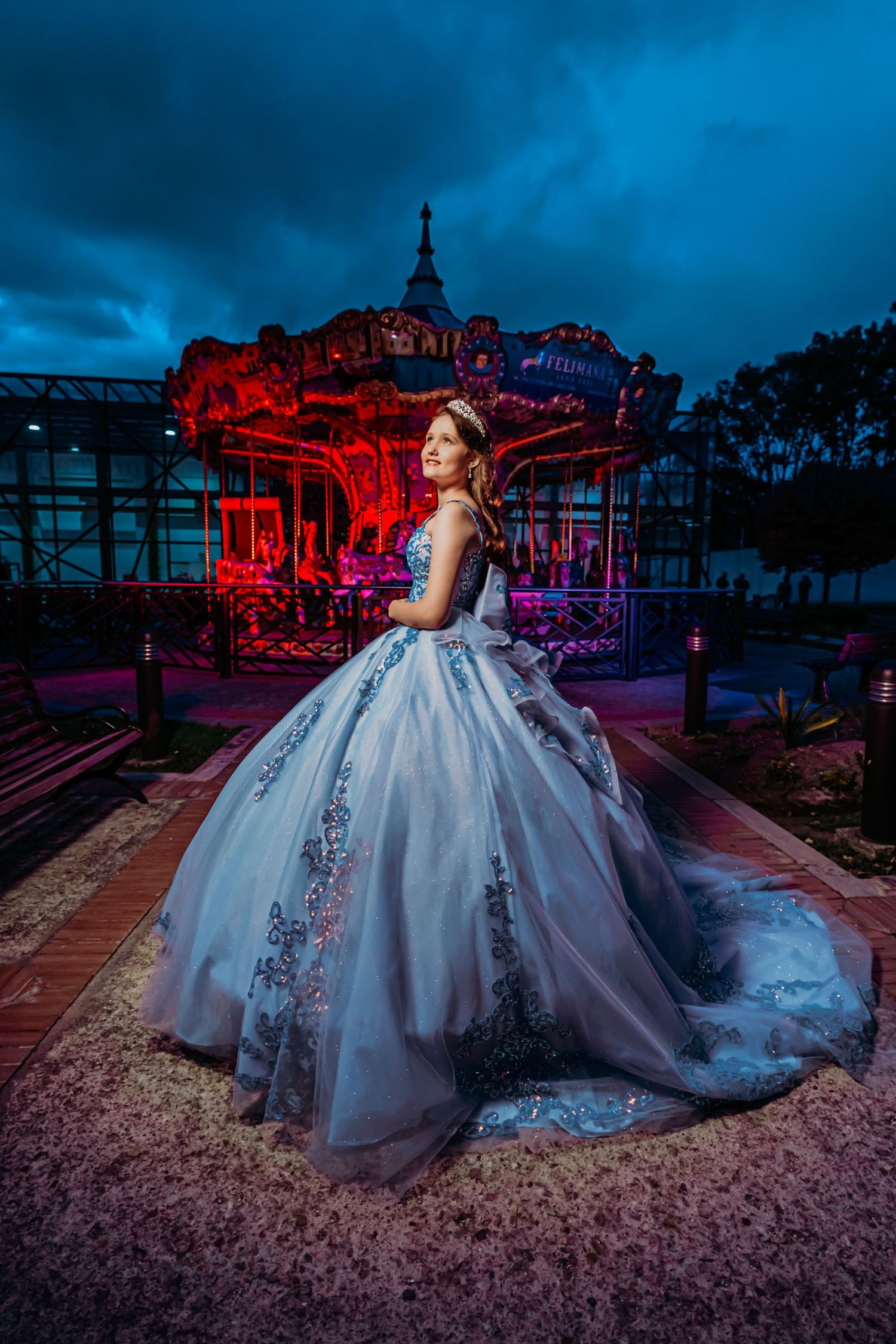 Quinceañera con vestido azul princesa y tiara posa de noche frente a carrusel iluminado rojo y azul.